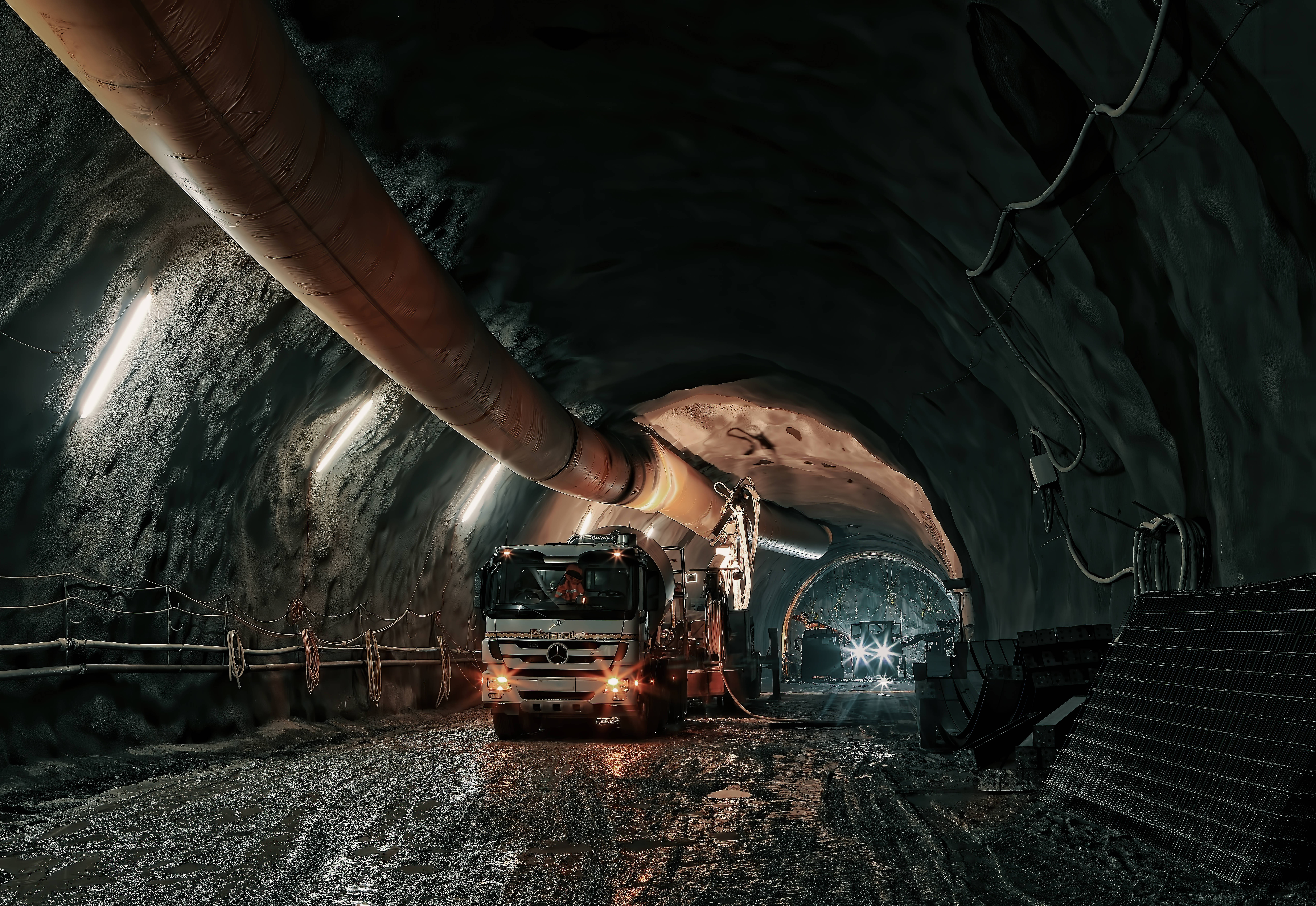 Mining 桌布 壁紙 — 一架重型礦用卡車在光線昏暗的地下隧道中行駛，隧道內有裸露的岩壁和頭頂的工業管道。