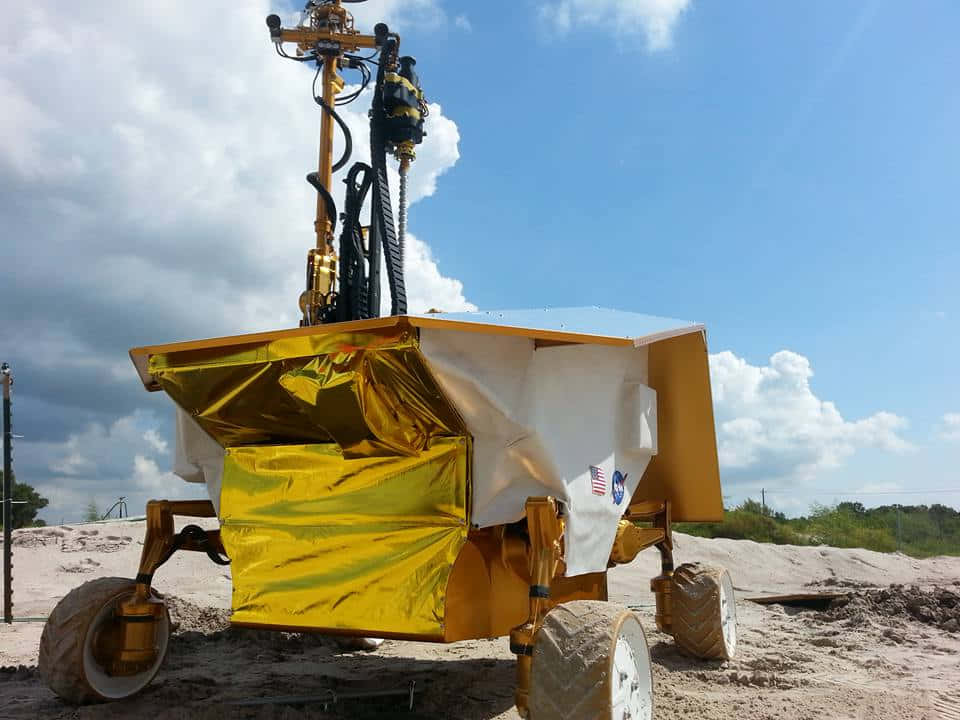 Lunar Rover 桌布 壁紙 — 一架金色和白色的NASA火星探測車站在沙質地形上，天空部分多雲，遠處有綠樹。