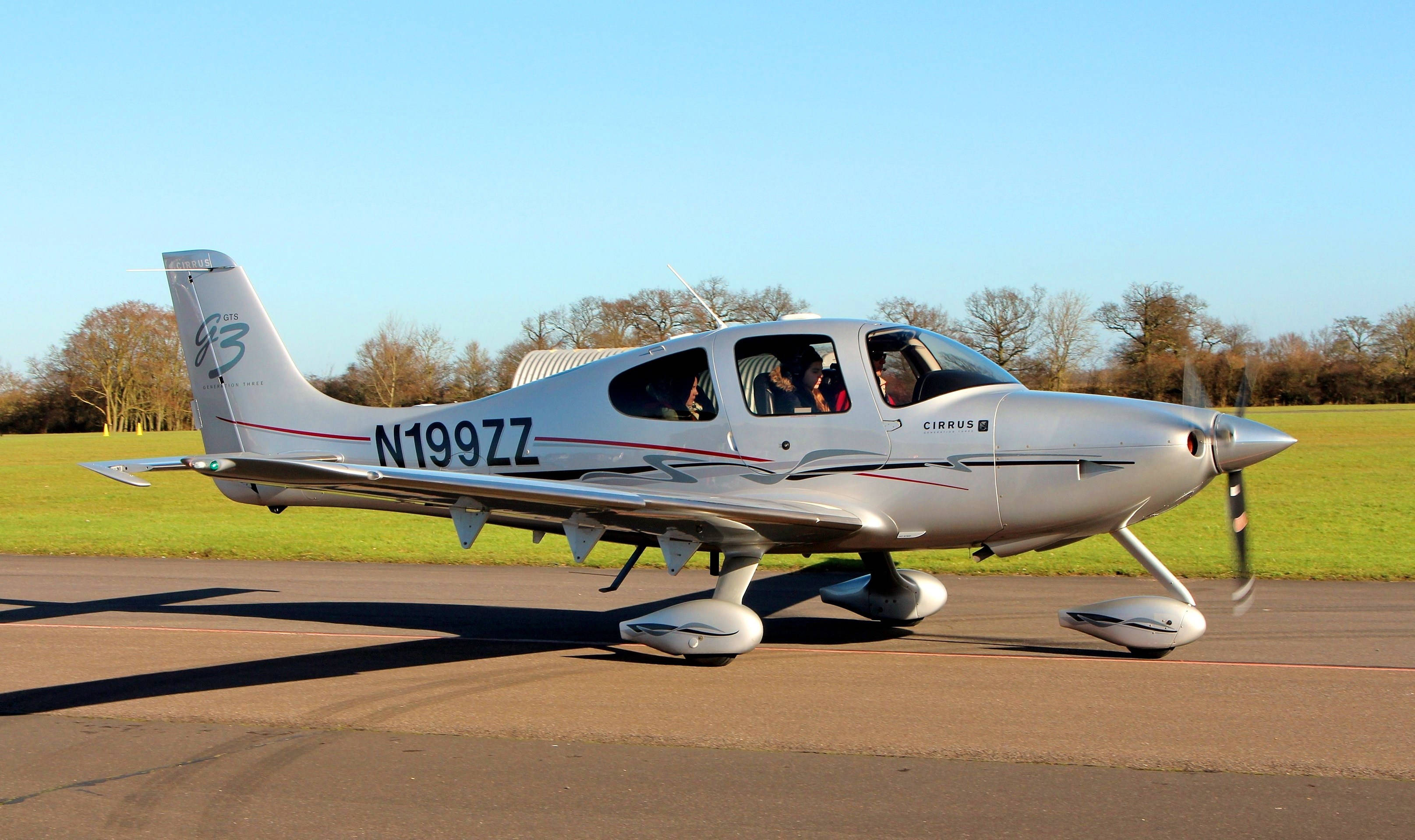 Single 桌布 壁紙 — 一架銀色 Cirrus SR22 飛機（註冊編號 N199ZZ）在晴朗藍天下、綠色田野和光禿樹木的背景中於跑道上滑行。
