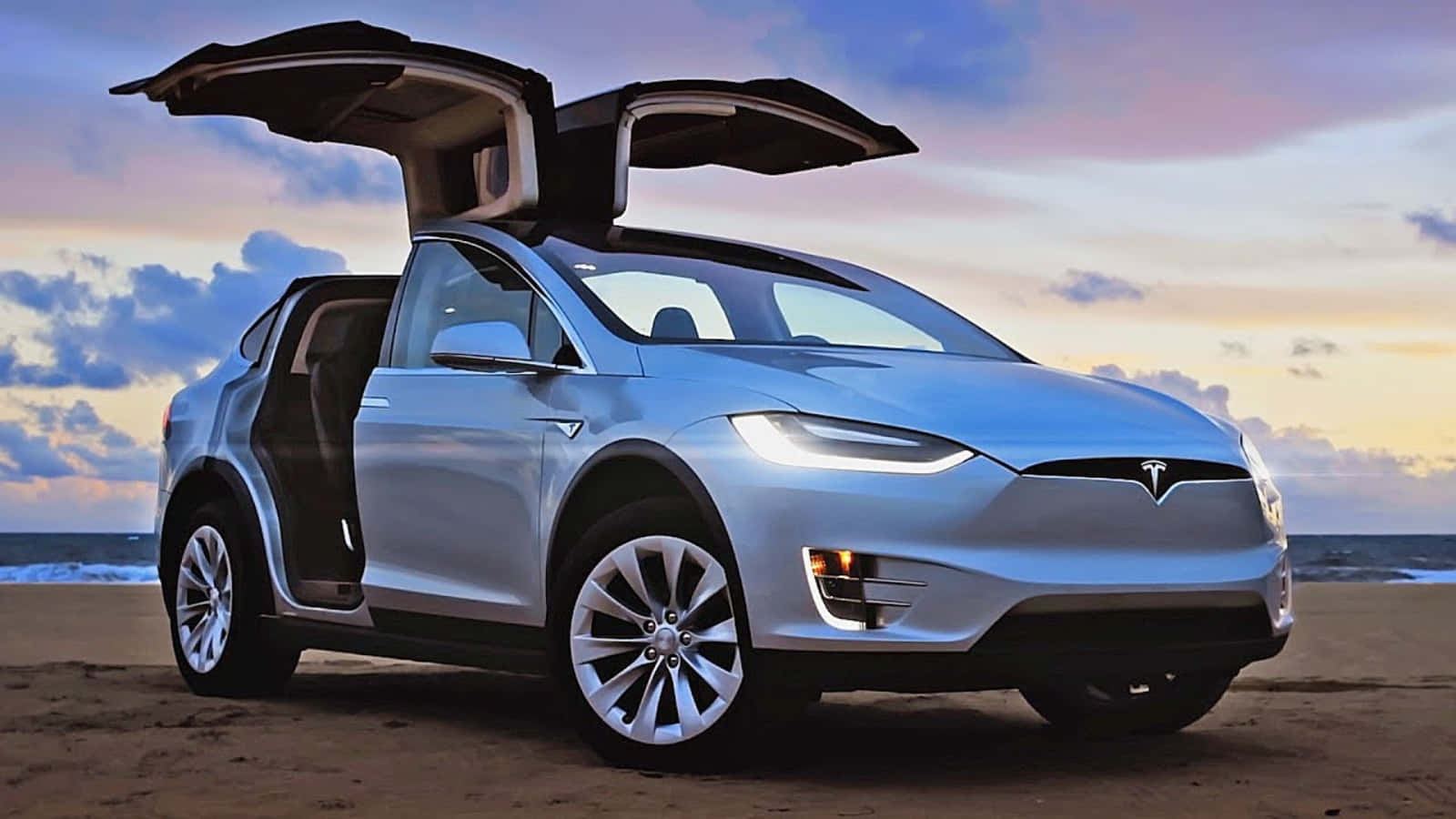 特斯拉 桌布 壁紙 — 一架銀色 Tesla Model X 打開鷗翼門，站在日落時分的沙灘上，背景有海浪。