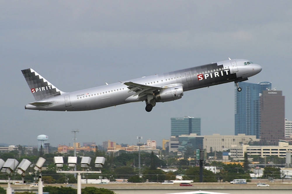 飛機 桌布 壁紙 — 一架銀色精神航空噴射機在陰雲密布的天空中攀升，下方是城市天際線和機場基礎設施。