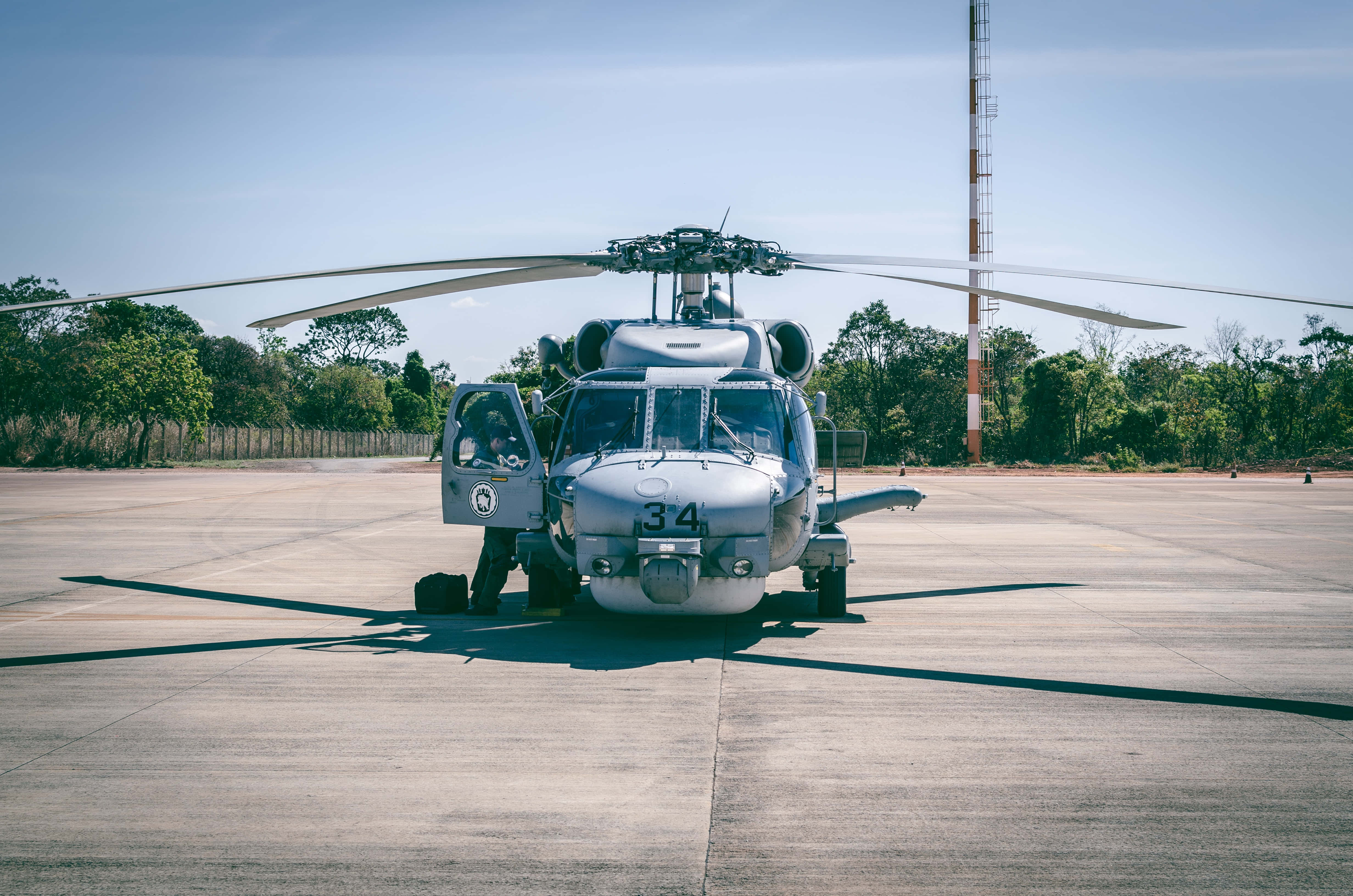Helicopter 桌布 壁紙 — 一架銀色軍用直升機，旋翼展開，停在混凝土停機坪上，背景是晴朗的藍天和樹木。