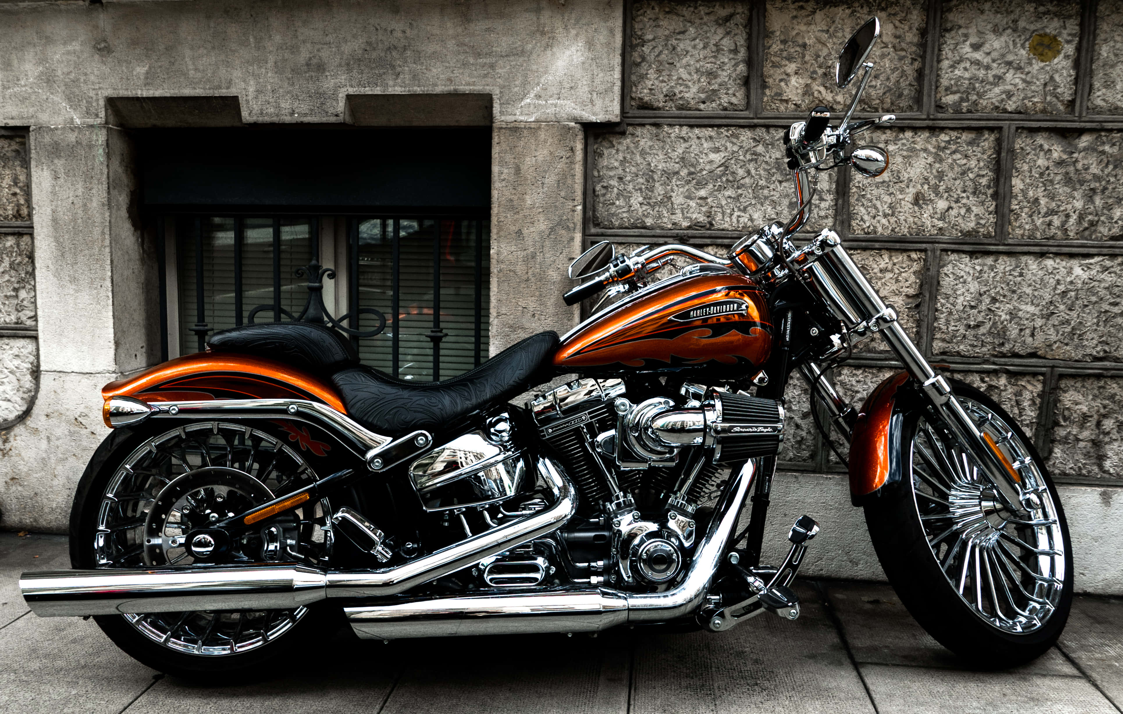Harley Davidson HD 桌布 壁紙 — 一架閃亮的橙色和鉻色哈雷戴維森摩托車停靠在風化的石牆旁。