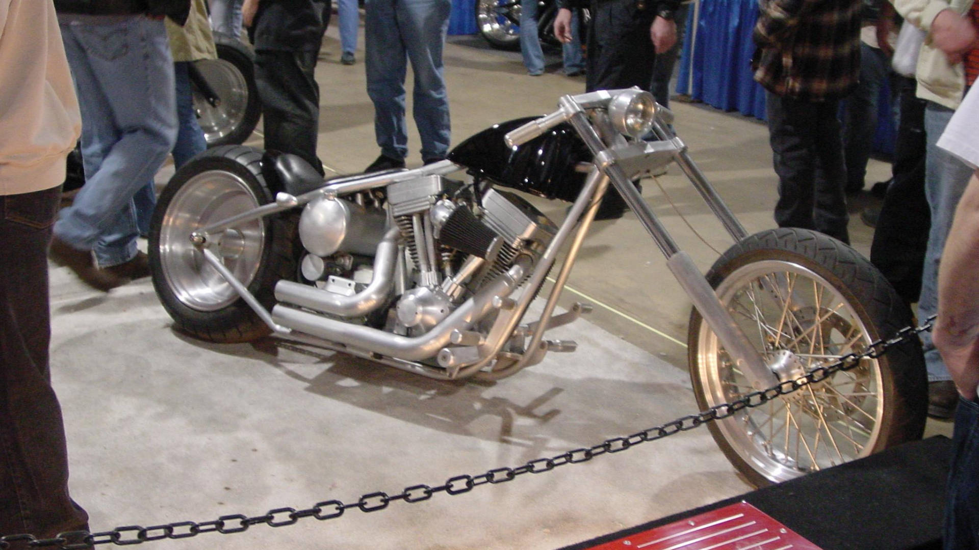 Choppers 桌布 壁紙 — 一架閃亮的訂製chopper機車，帶有鍍鉻細節和輻條輪，在室內展覽中展出，周圍有觀眾。