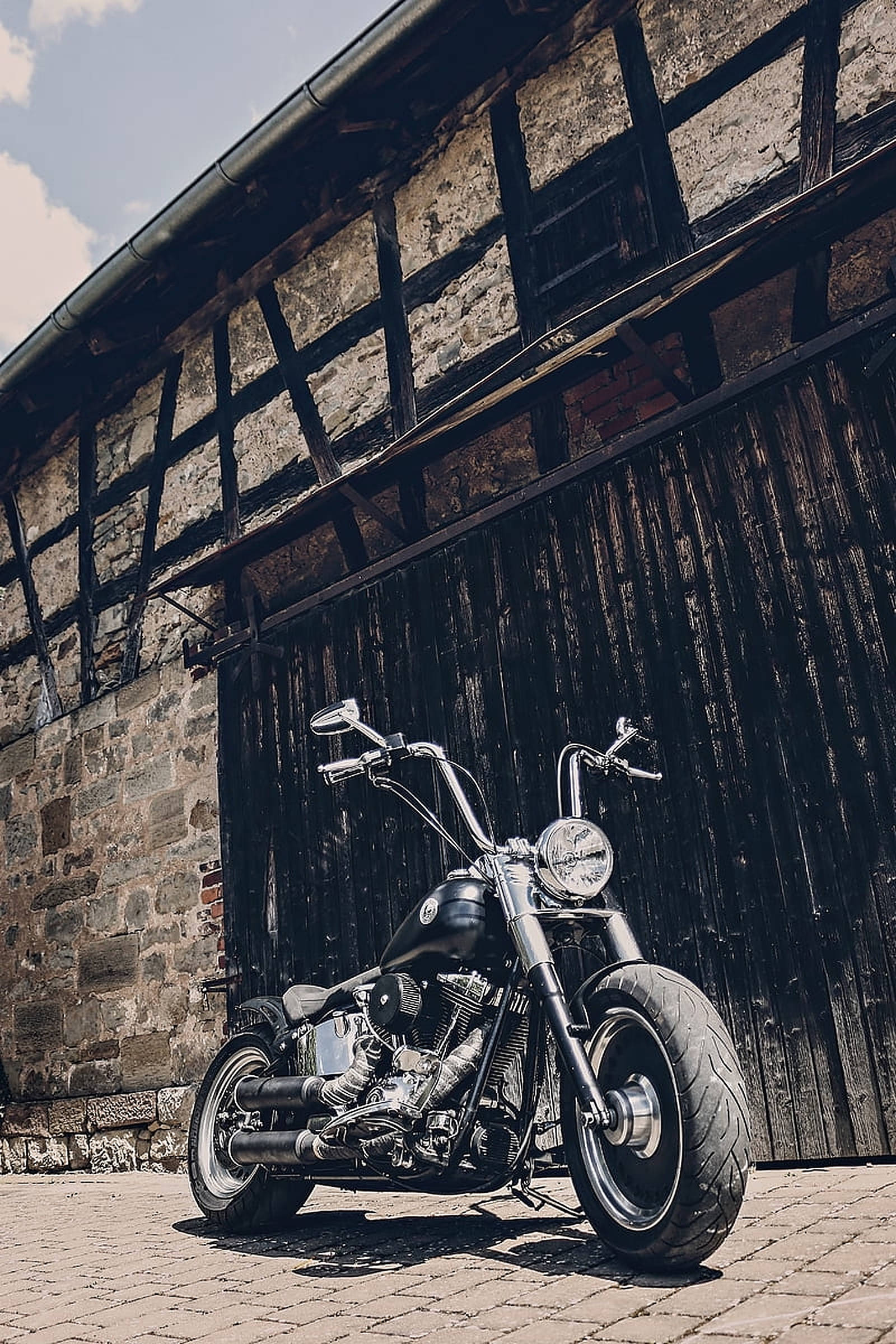 Chopper 桌布 壁紙 — 一架閃亮的黑色chopper機車停泊在鵝卵石路上，前方是質樸的半木造石建築，天空明亮。