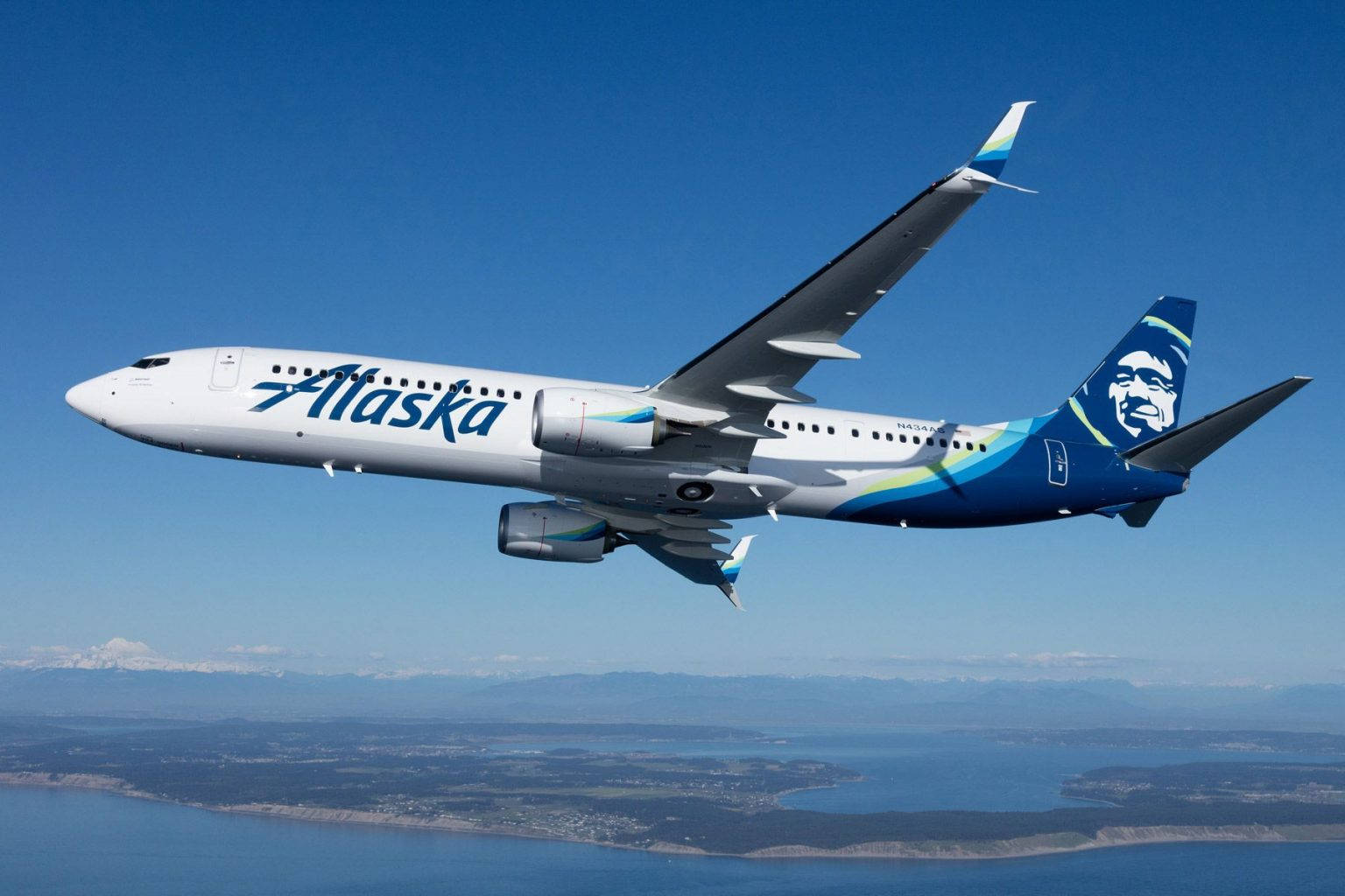 飛機 桌布 壁紙 — 一架阿拉斯加航空的飛機在晴朗的藍天下翱翔於海岸景觀之上，遠方有積雪覆蓋的山脈。