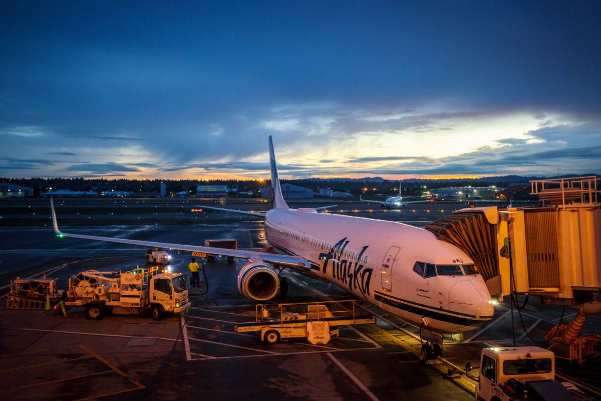 飛機 桌布 壁紙 — 一架阿拉斯加航空的飛機在黃昏時停靠在機場閘口，周圍有地勤人員和設備，天空壯麗。