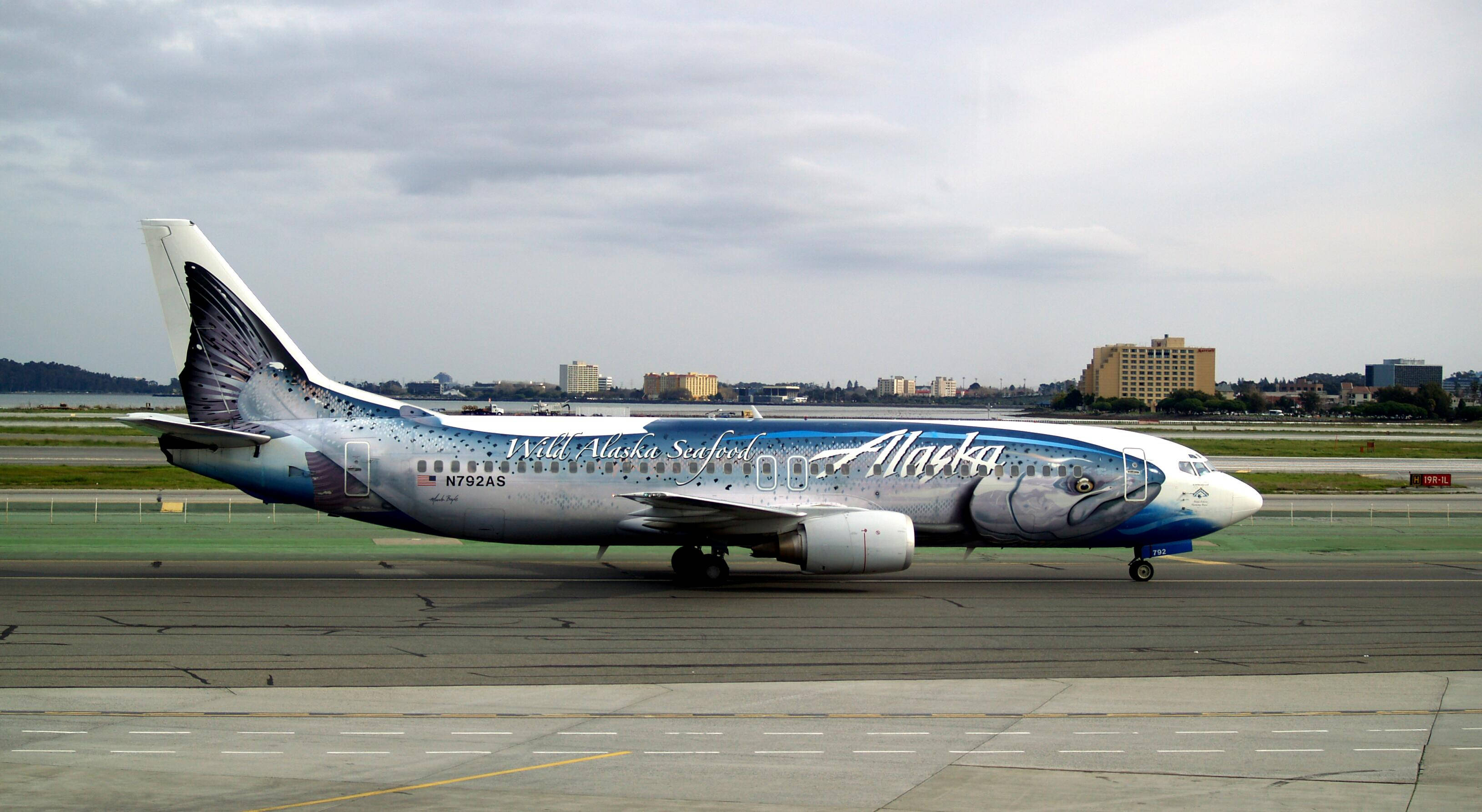 飛機 桌布 壁紙 — 一架阿拉斯加航空的飛機帶有野生鮭魚塗裝，在潮濕的跑道上滑行，天空多雲，遠處有城市建築。