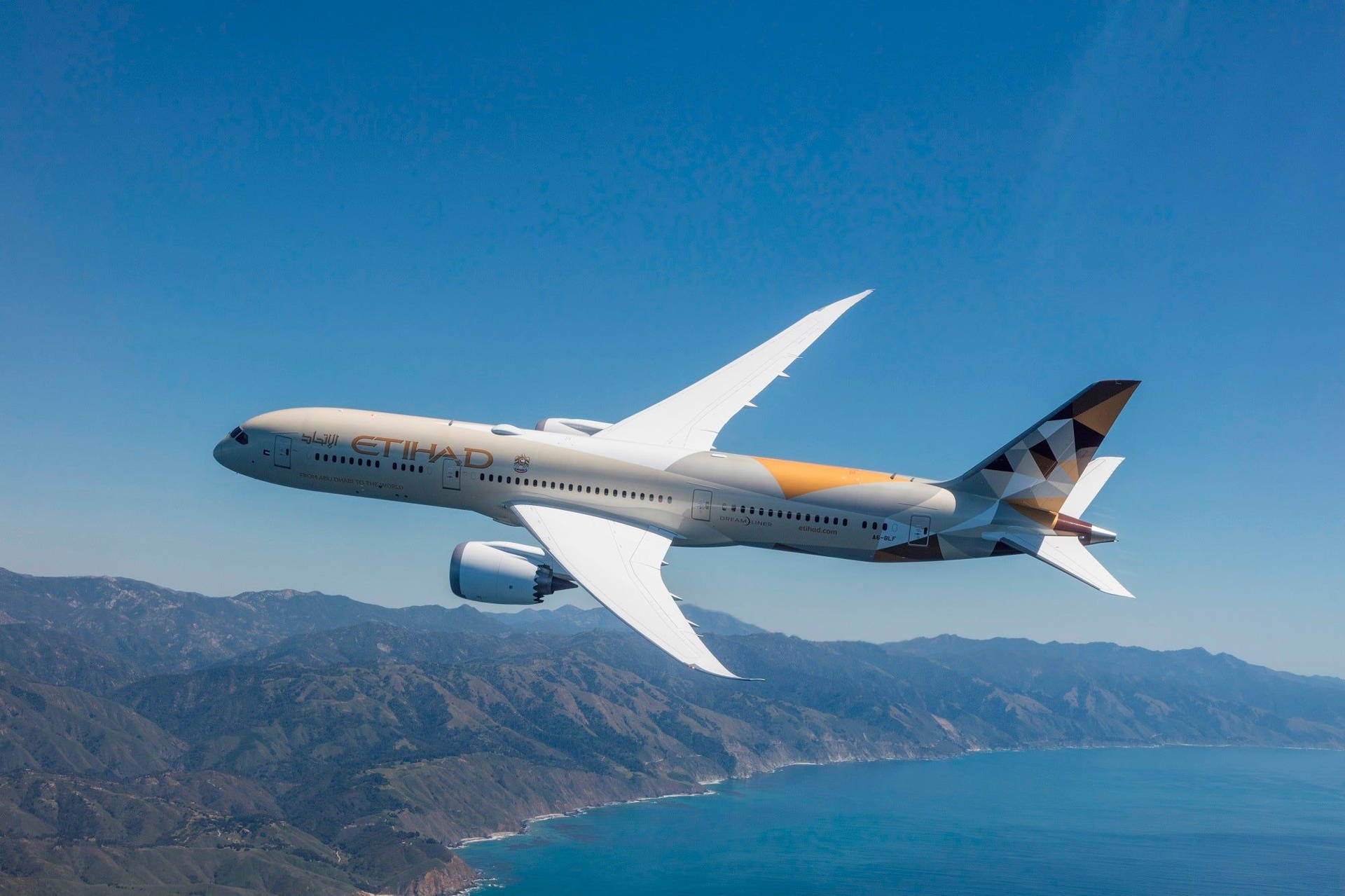  壁紙 — 一架阿提哈德航空的飛機在晴朗藍天下優雅地翱翔於沿海山脈之上。