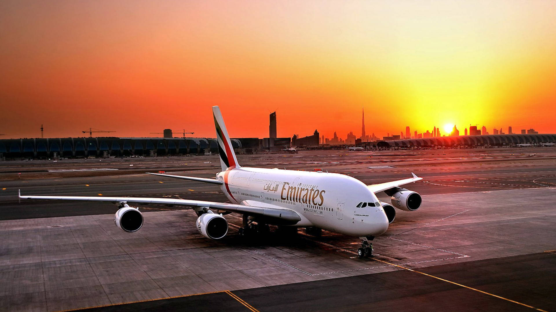  壁紙 — 一架阿聯酋航空的空中巴士A380停在機場停機坪上，夕陽在現代城市天際線後落下。