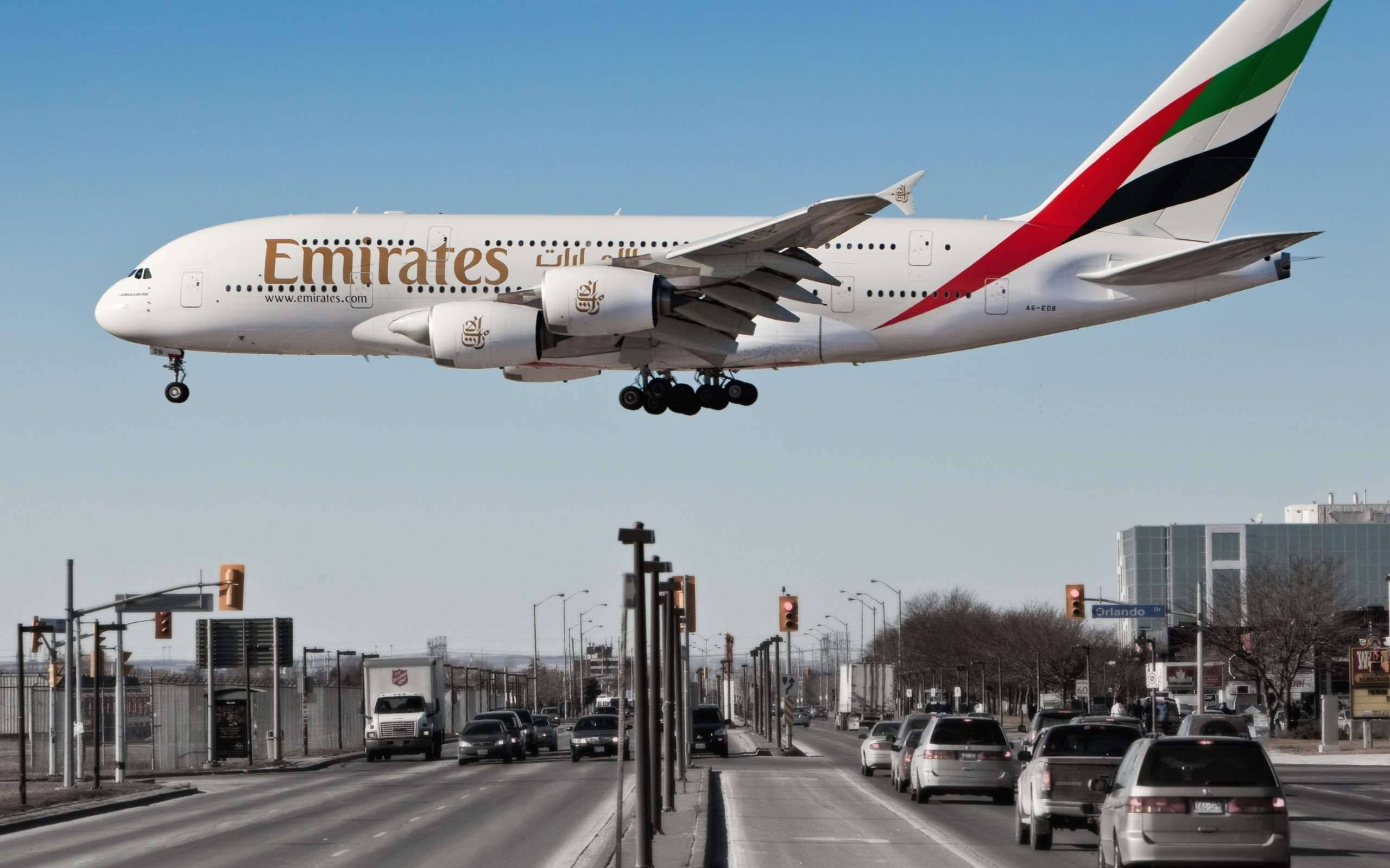 飛機 桌布 壁紙 — 一架阿聯酋航空空中巴士A380低飛過繁忙的城市高速公路，下方有汽車和交通號誌。