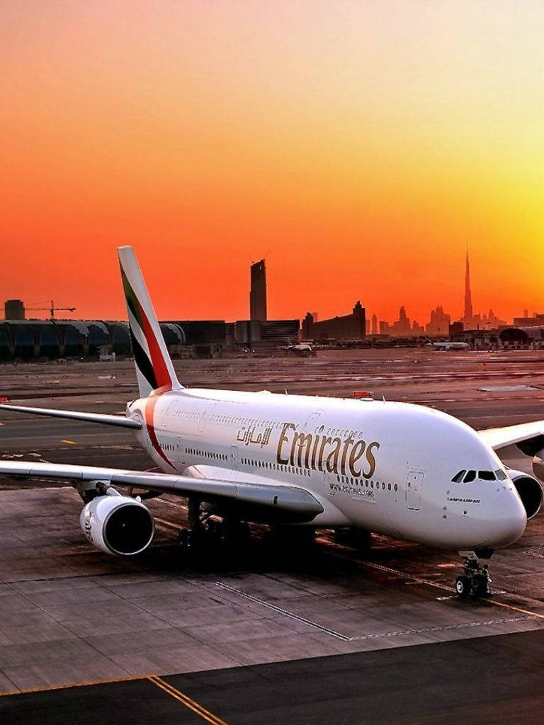 飛機 桌布 壁紙 — 一架阿聯酋航空空中巴士A380在日落時停泊在停機坪上，背後杜拜的天際線閃耀著橙色光芒。