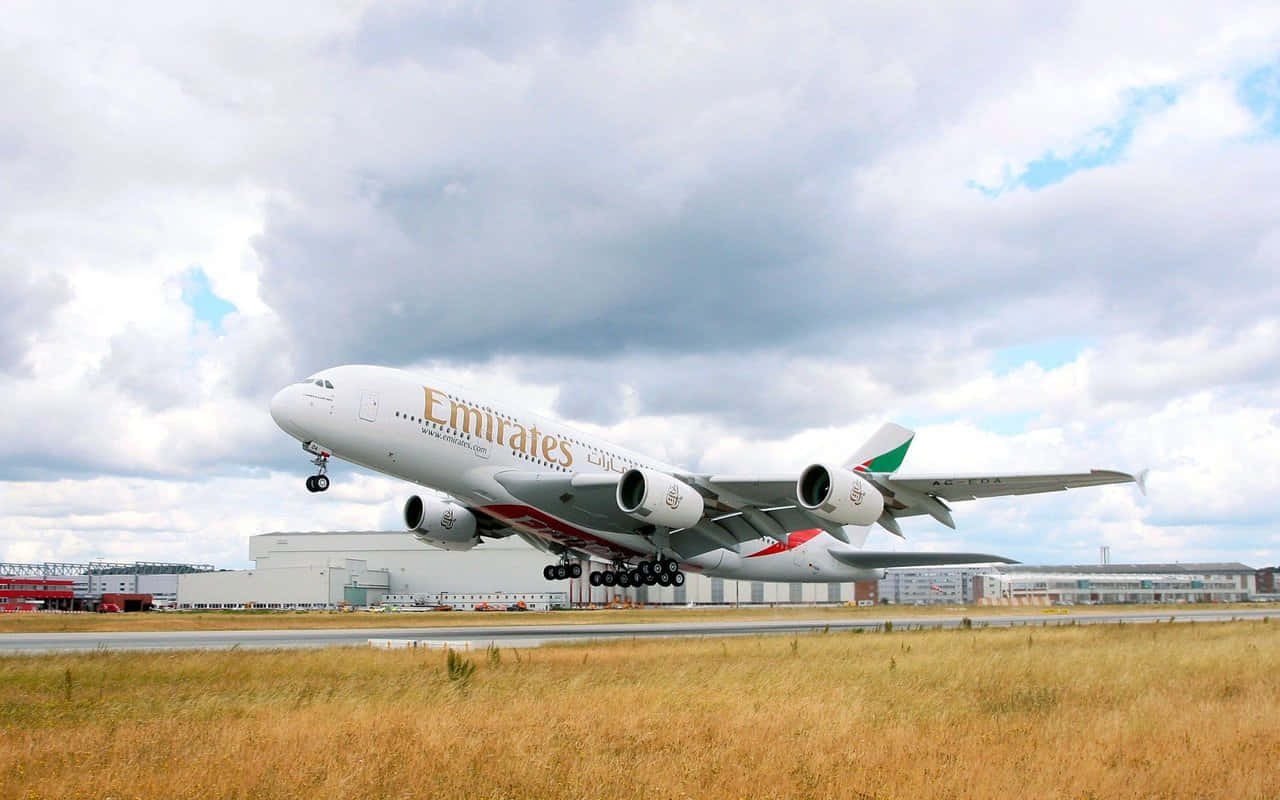 飛機 桌布 壁紙 — 一架阿聯酋航空空中巴士A380在多雲的天空下從機場跑道起飛，前景有金色的草。