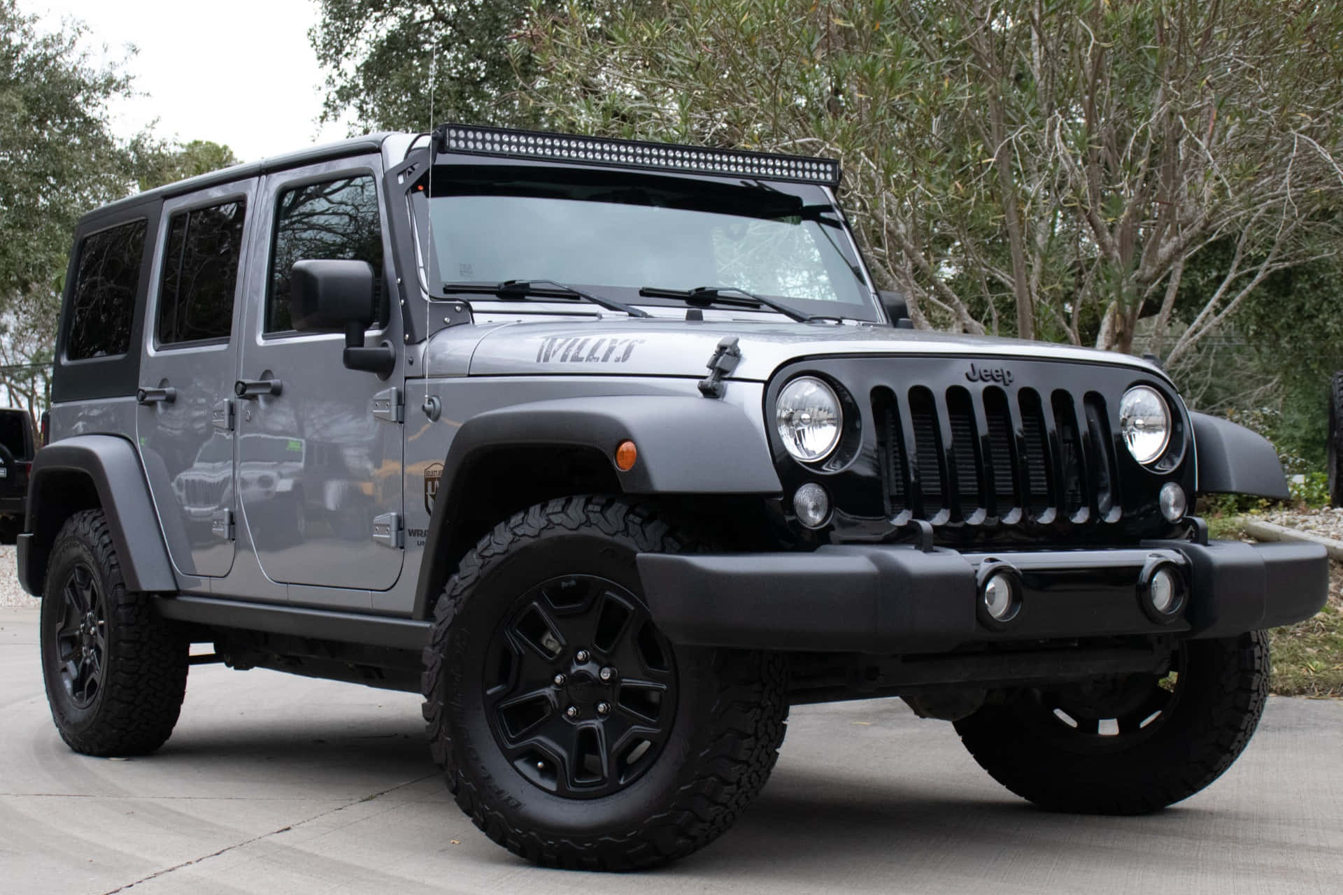 Jeep Willys 桌布 壁紙 — 一架霧面灰色Jeep Wrangler，帶有黑色飾件和越野輪胎，停放在樹木環繞的混凝土車道上。