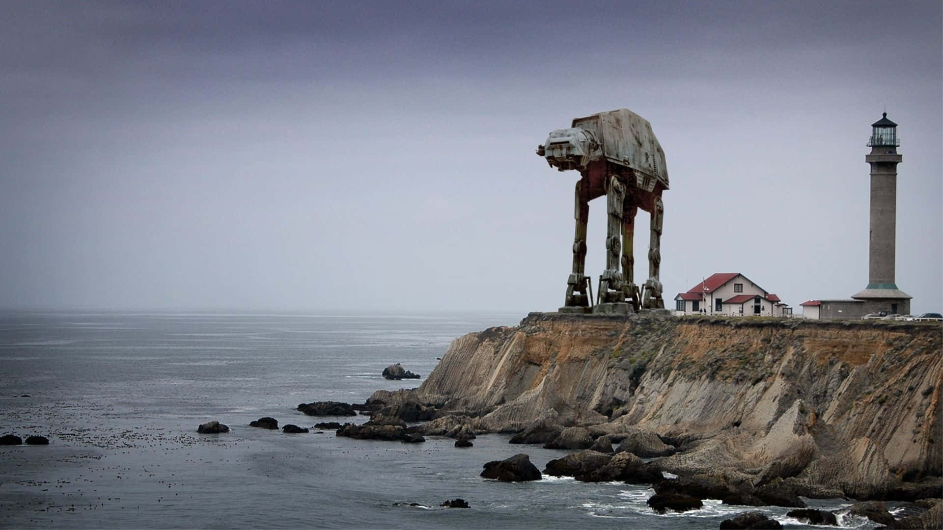 At-at 桌布 壁紙 — 一架風化的AT-AT步行者威嚴地矗立在岩石懸崖上，旁邊是燈塔和沿海建築，天空灰濛濛。