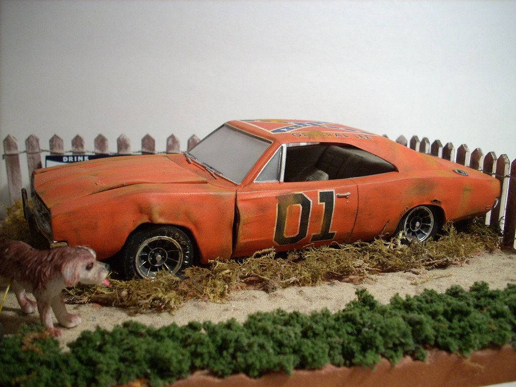 Dukes Of Hazzard 桌布 壁紙 — 一架風化的橘色 Dodge Charger 模型，側面有「01」字樣，放在乾草上，旁邊有木柵欄和一個小狗公仔。