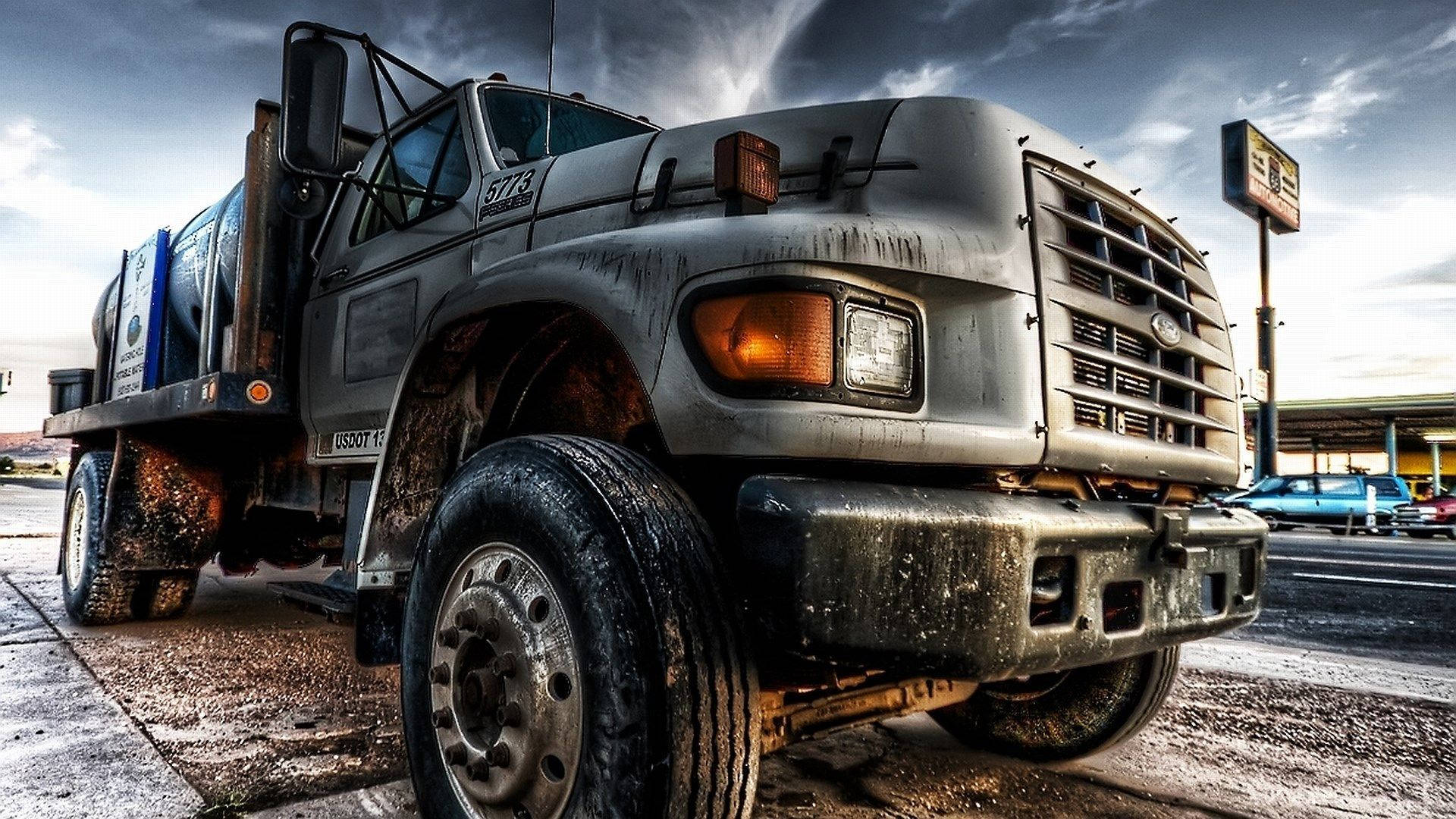 Trucks 桌布 壁紙 — 一架風化的灰色自卸車，輪胎沾滿泥濘，停在一個加油站，天空烏雲密布，氣氛戲劇化。