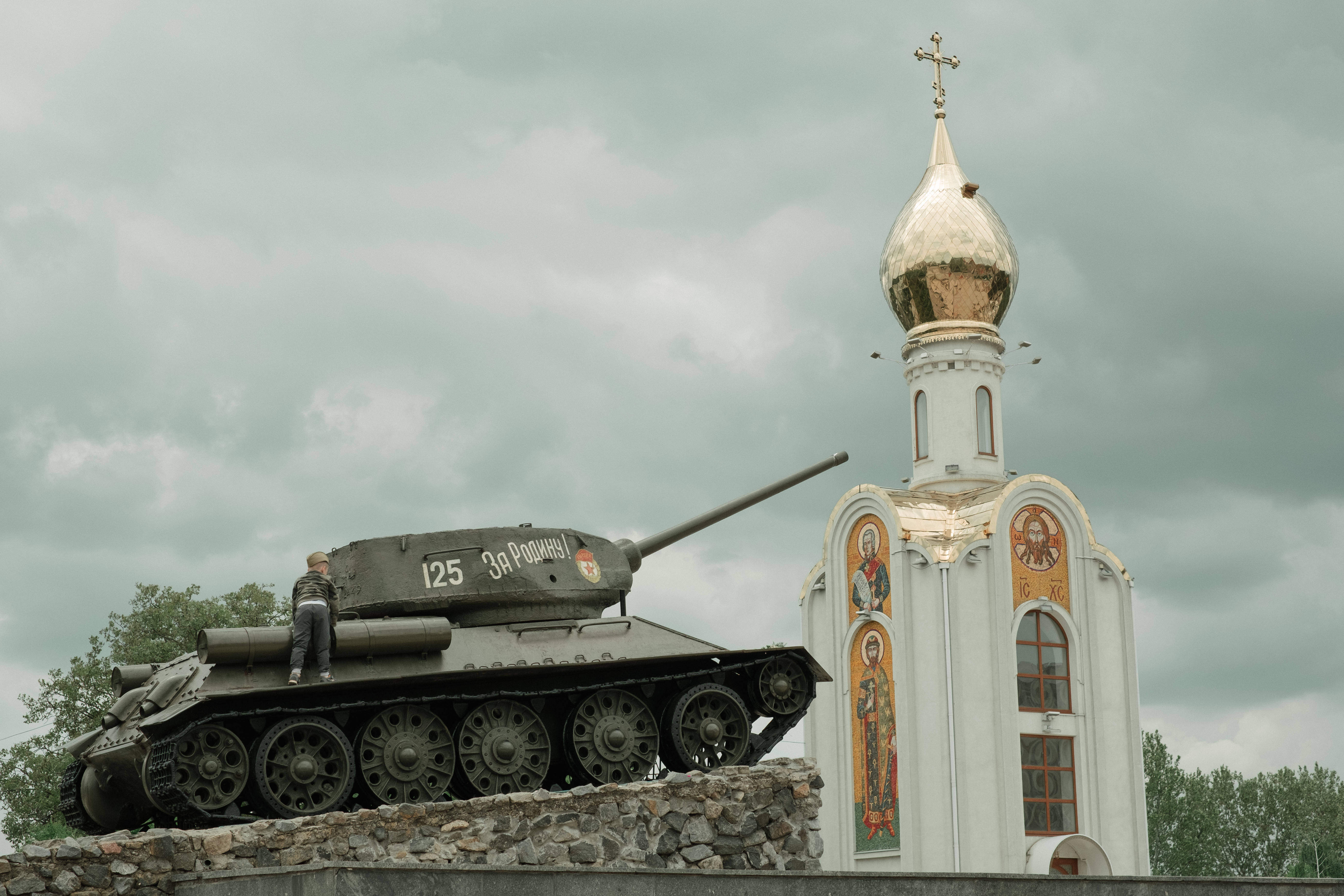 Moldova 桌布 壁紙 — 一架風化的軍用坦克，旁邊站著一名士兵，旁邊是一座白色東正教教堂，金色圓頂，天空多雲。