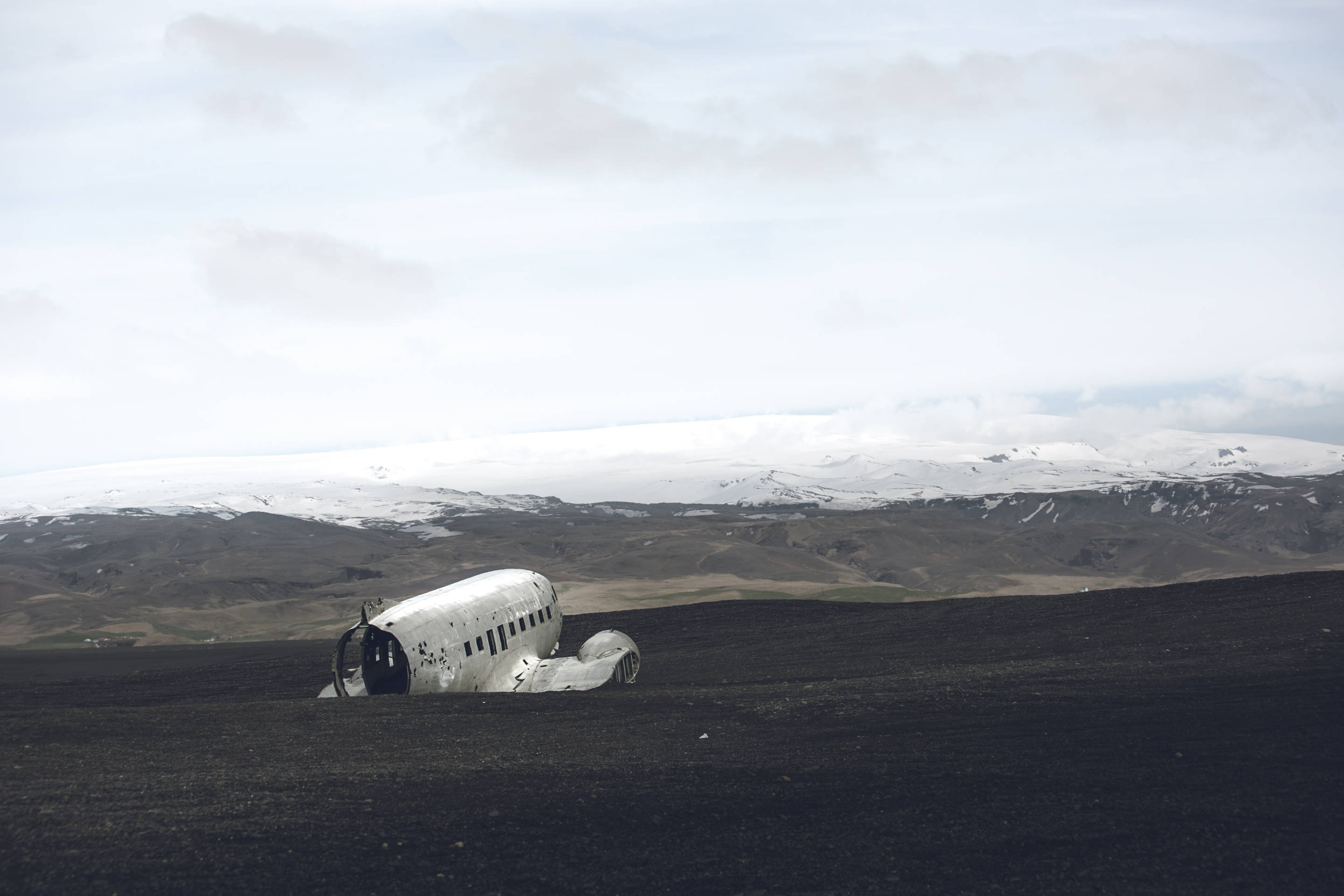 Plane 桌布 壁紙 — 一架風化的飛機機身停放在黑色火山岩地上，遠方有積雪的山脈，天空多雲。