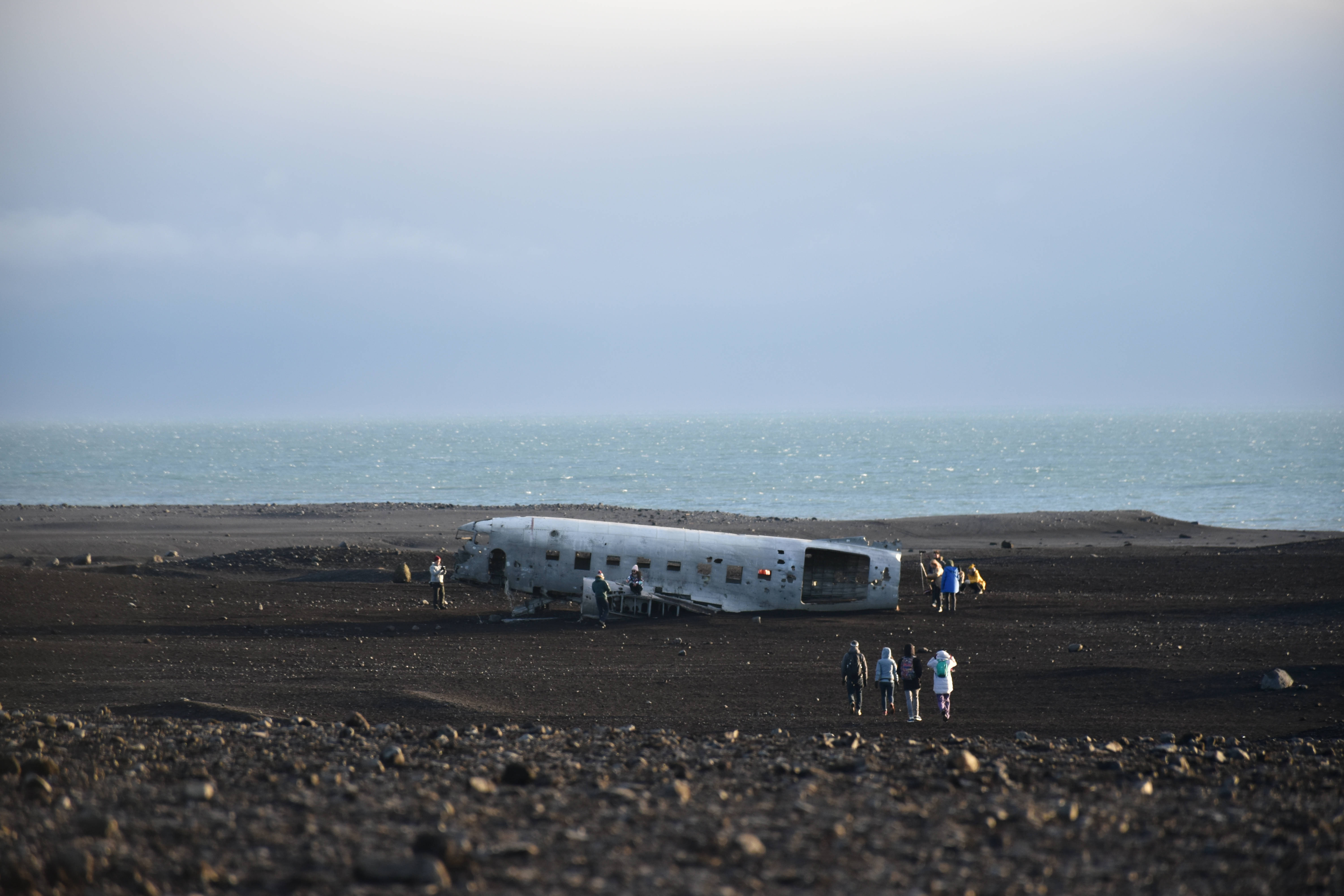 Plane 桌布 壁紙 — 一架風化的飛機機身擱在黑色火山海灘上，遊客正在海洋附近探索殘骸。