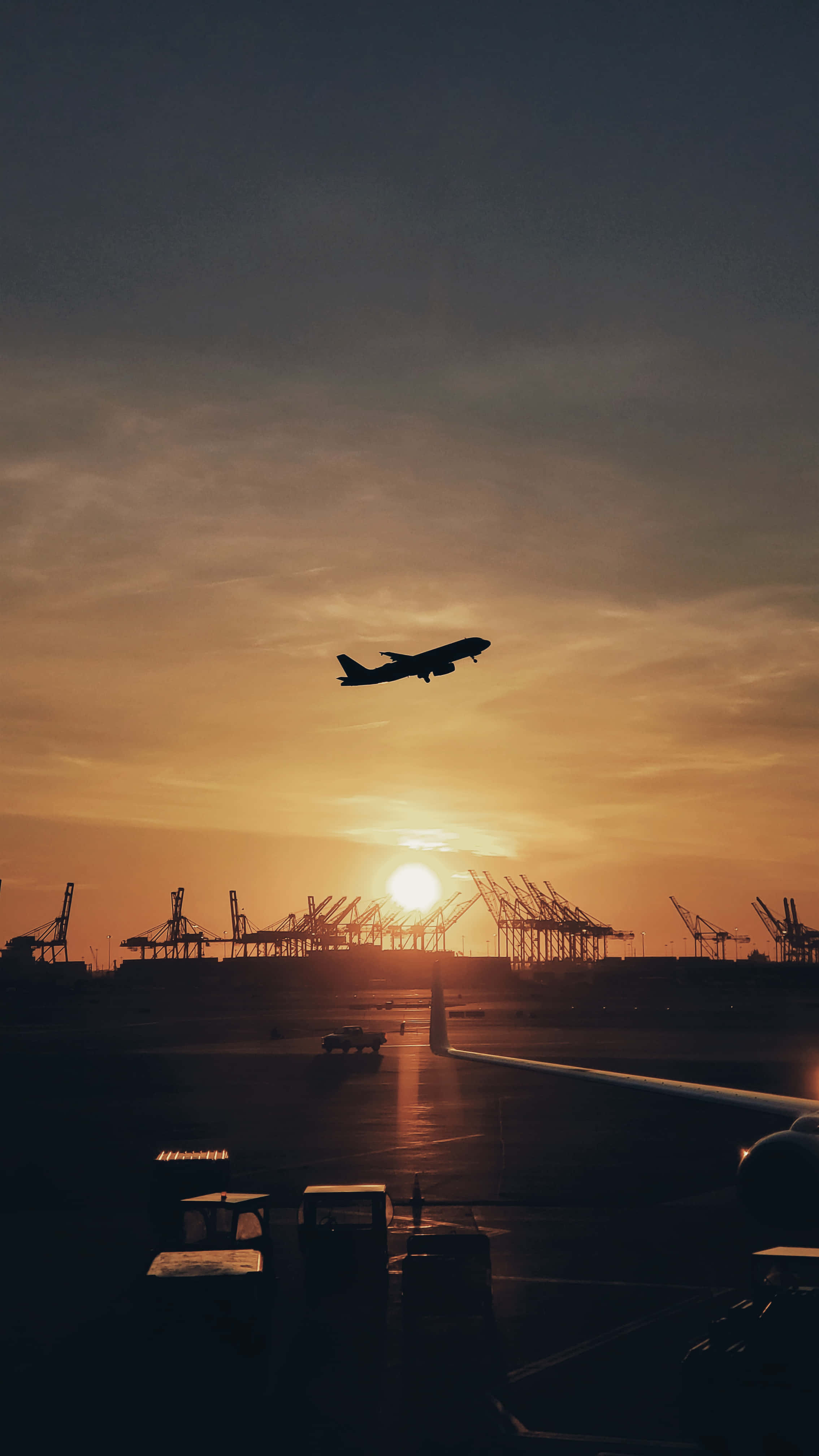 飛機 桌布 壁紙 — 一架飛機在金色夕陽的映襯下飛過機場，遠處有起重機。