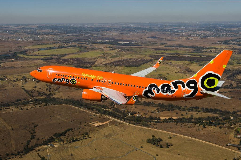 飛機 桌布 壁紙 — 一架鮮橙色的Mango Airlines飛機在晴朗天空下，翱翔於乾旱田野與遠處城鎮的拼布之上。