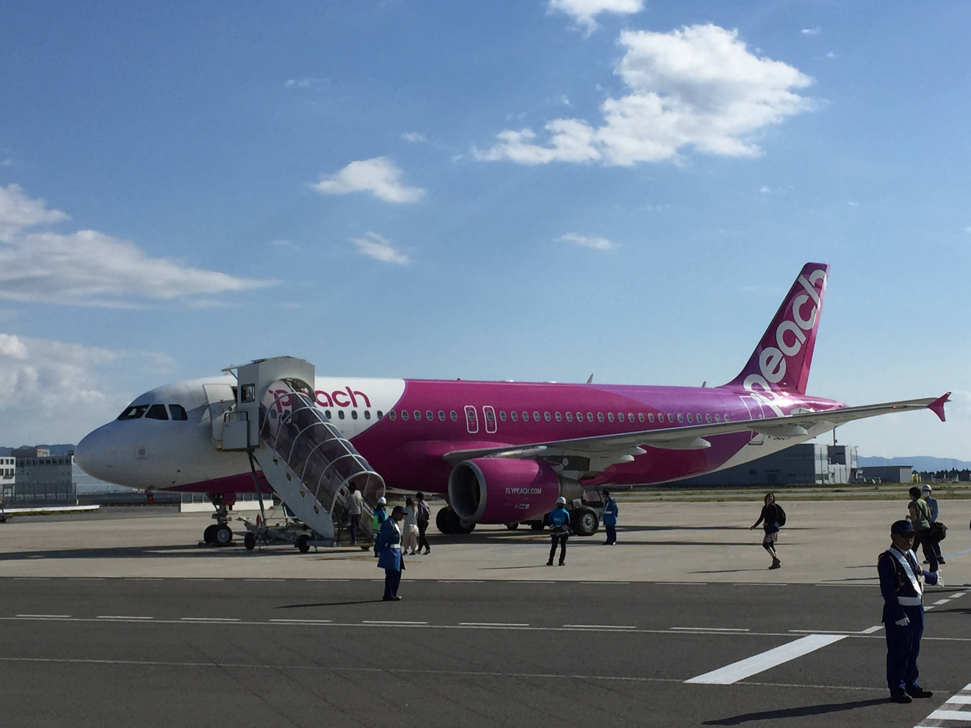 Airport 桌布 壁紙 — 一架鮮粉紅色的Peach Air飛機停在陽光明媚的停機坪上，乘客正透過登機橋登機，地勤人員在附近。