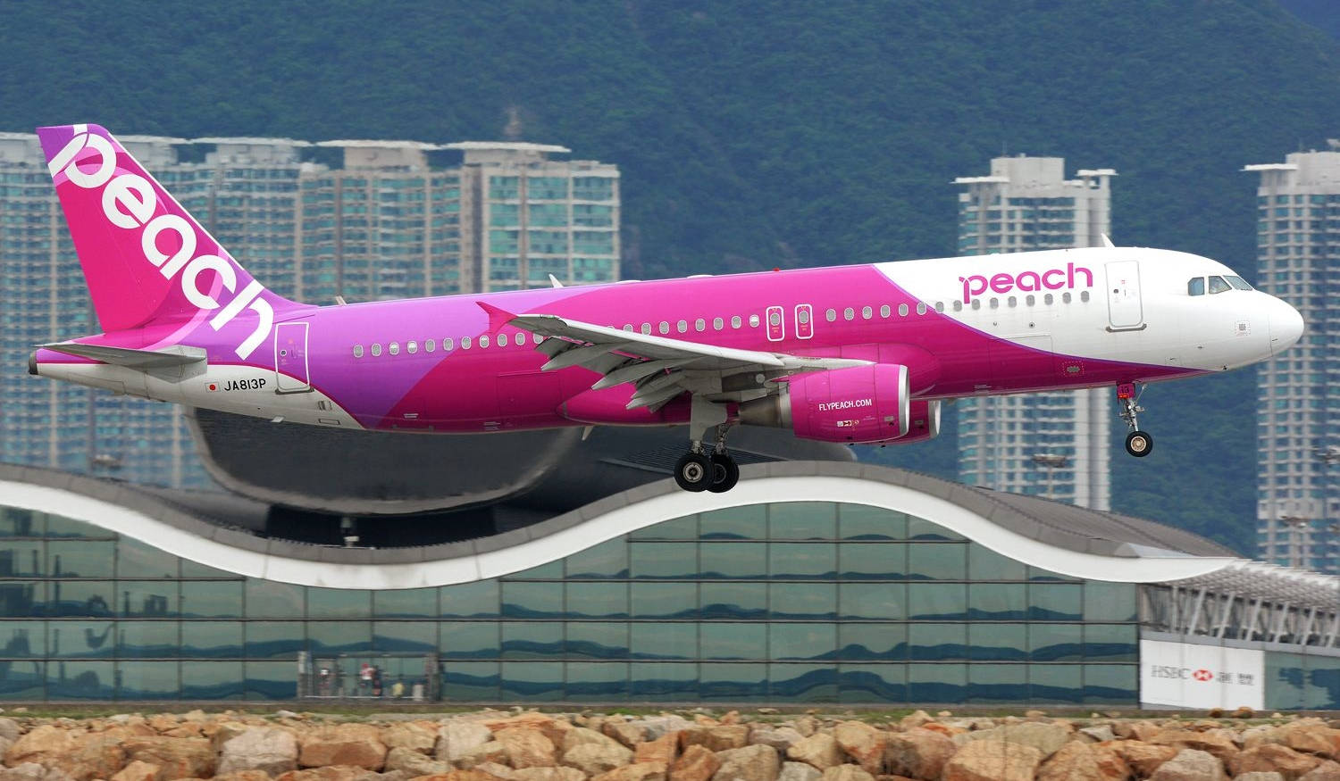 Airport 桌布 壁紙 — 一架鮮粉紅色的Peach Airlines飛機在現代摩天大樓和綠色山丘的背景下起飛。