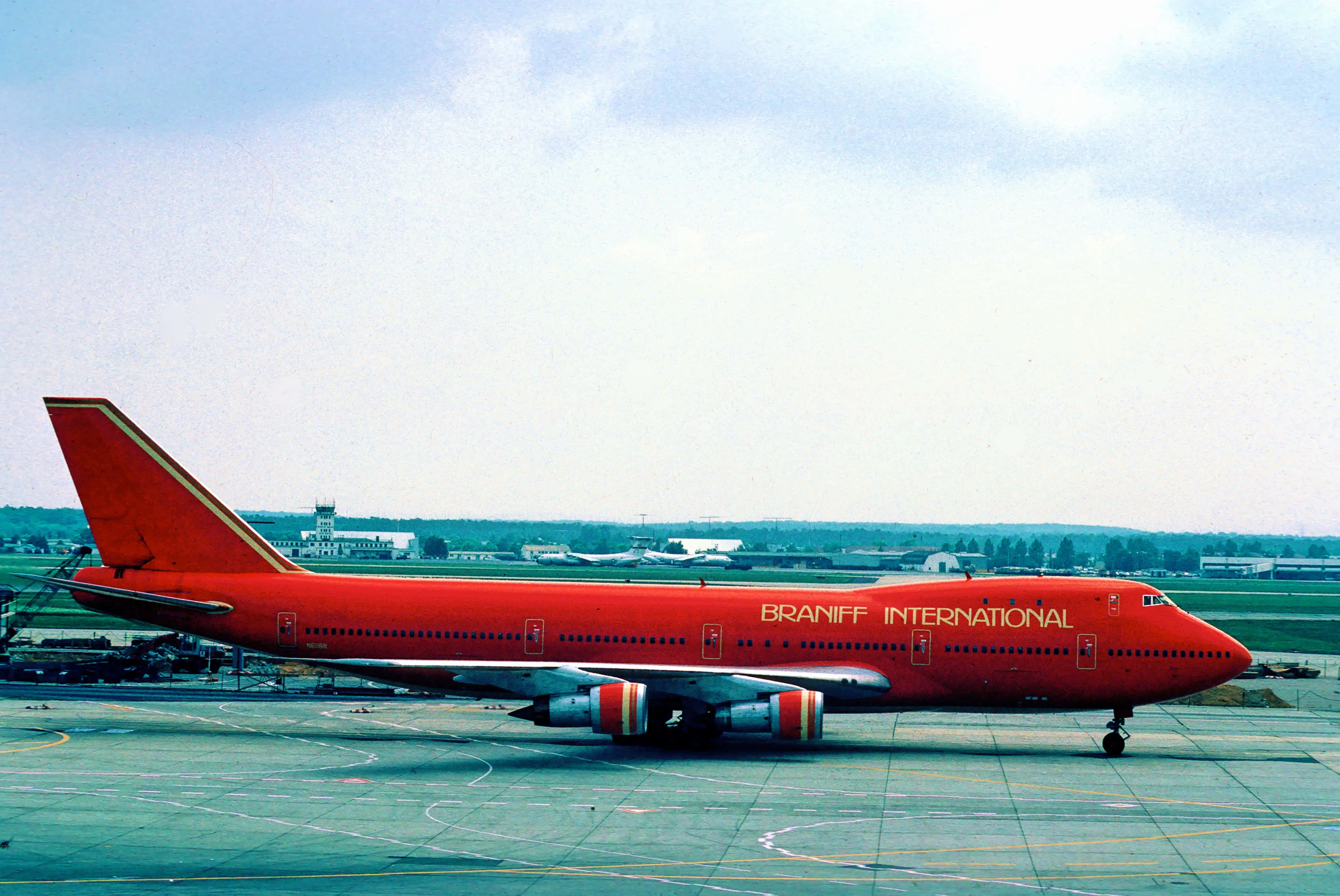 Jumbo Jets 桌布 壁紙 — 一架鮮紅色的 Braniff International 波音 747 停放在機場停機坪上，天空多雲，遠處有機棚和跑道標記。