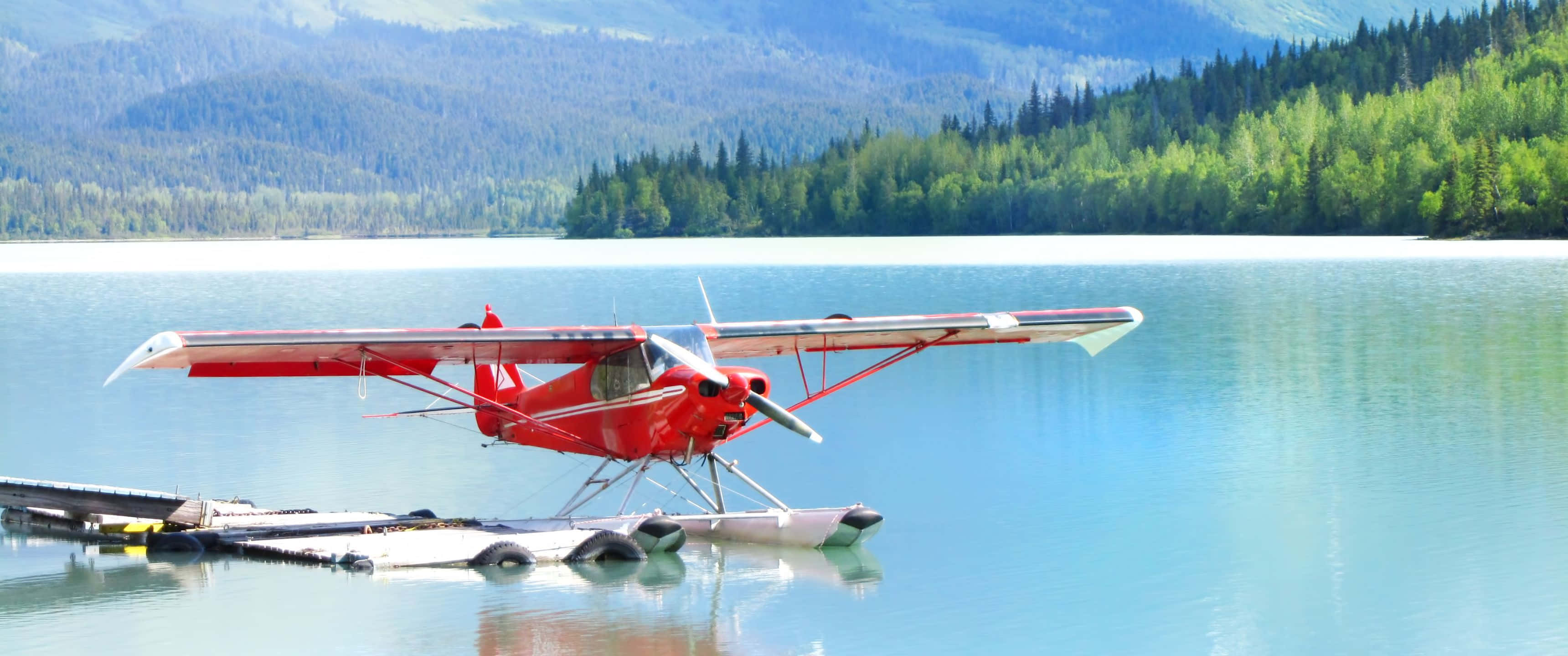3440x1440p Small Planes 桌布 壁紙 — 一架鮮紅色的水上飛機停泊在平靜的碧綠水面上，旁邊是木製碼頭，背景是樹木茂密的山巒。
