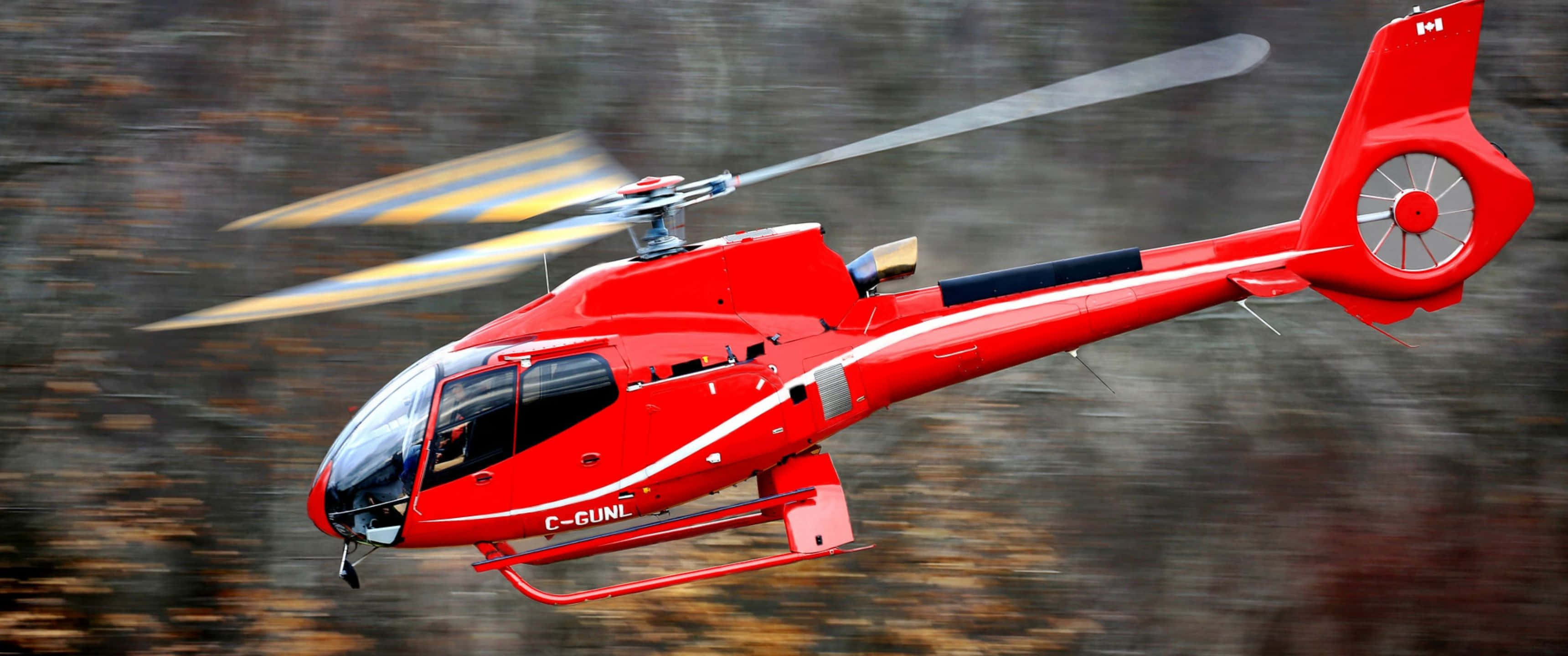 3440x1440p Helicopters 桌布 壁紙 — 一架鮮紅色的直升機，帶有模糊的旋翼葉片，在森林背景中動態飛翔。