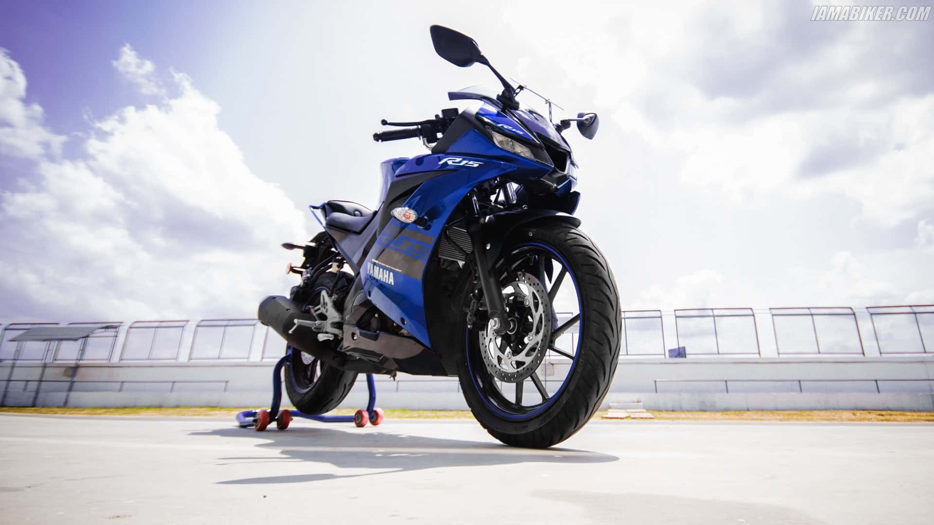 R15 桌布 壁紙 — 一架鮮藍色的Yamaha R15機車在晴朗天空和零星雲朵下立於支架上。