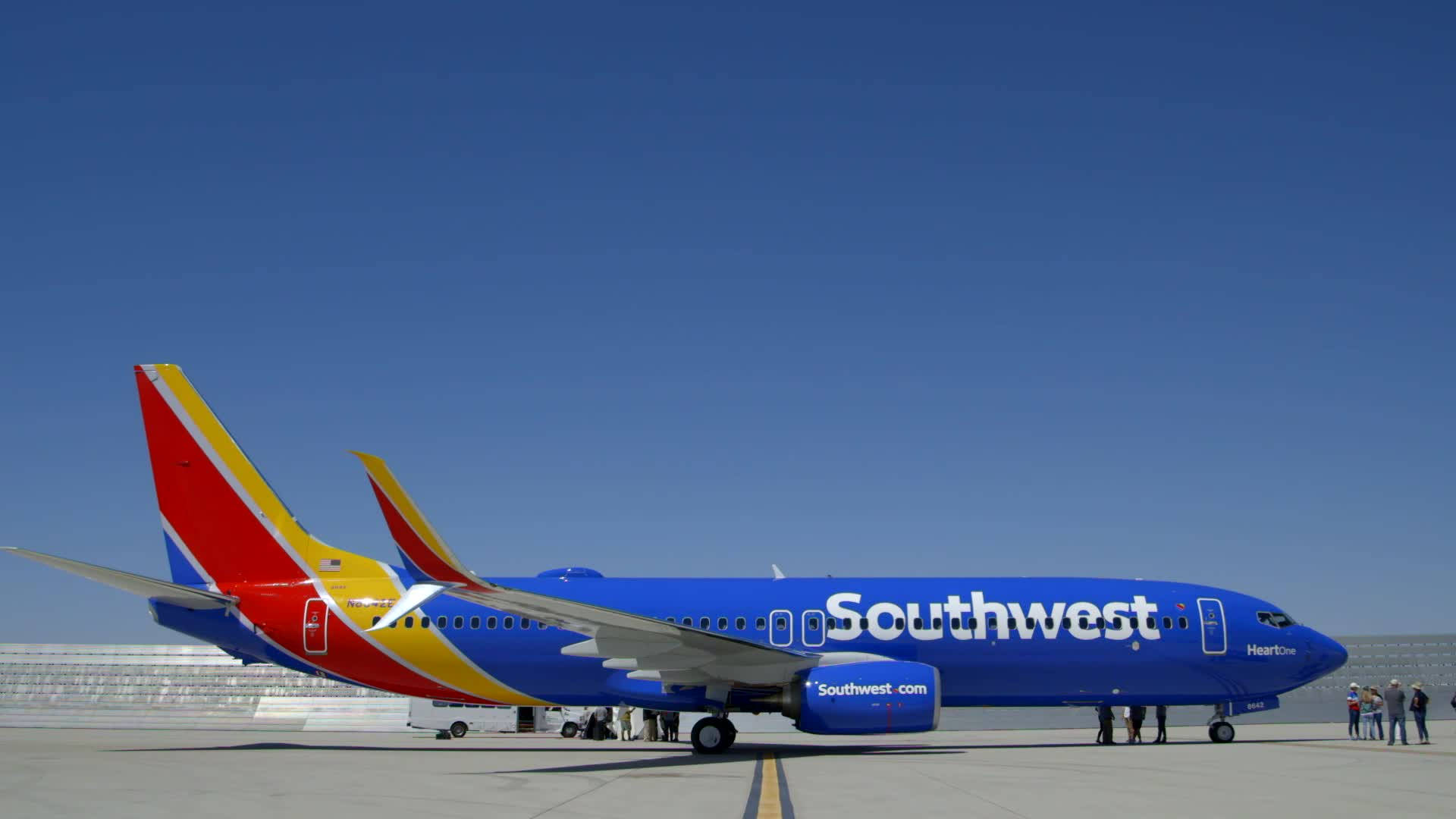 Southwest 桌布 壁紙 — 一架鮮藍色的西南航空飛機，帶有紅黃色尾翼，停在晴朗天空下的陽光停機坪上。