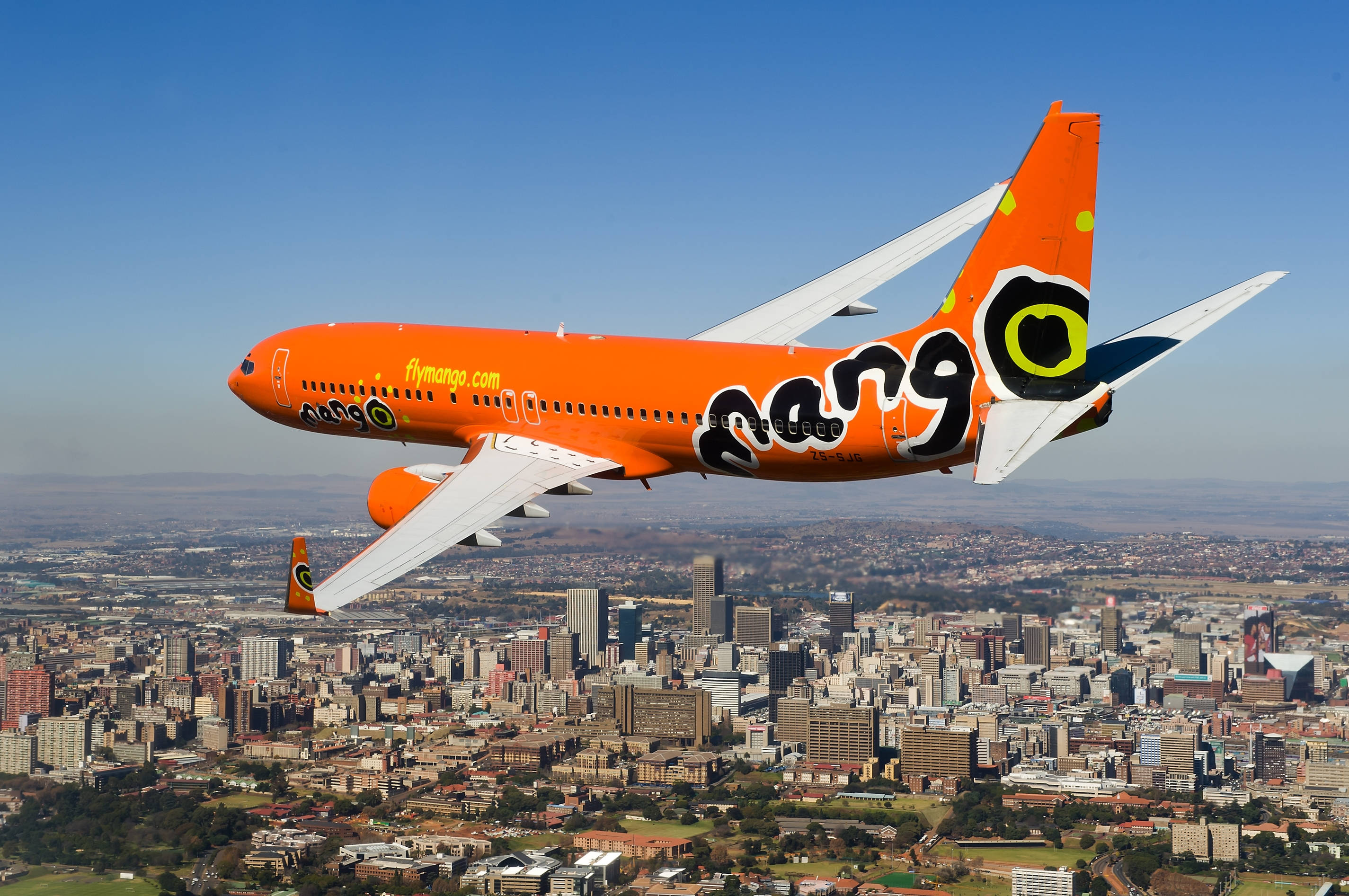 飛機 桌布 壁紙 — 一架鮮豔的橙色Mango Airlines飛機在晴朗的藍天下翱翔於廣闊的城市景觀之上。