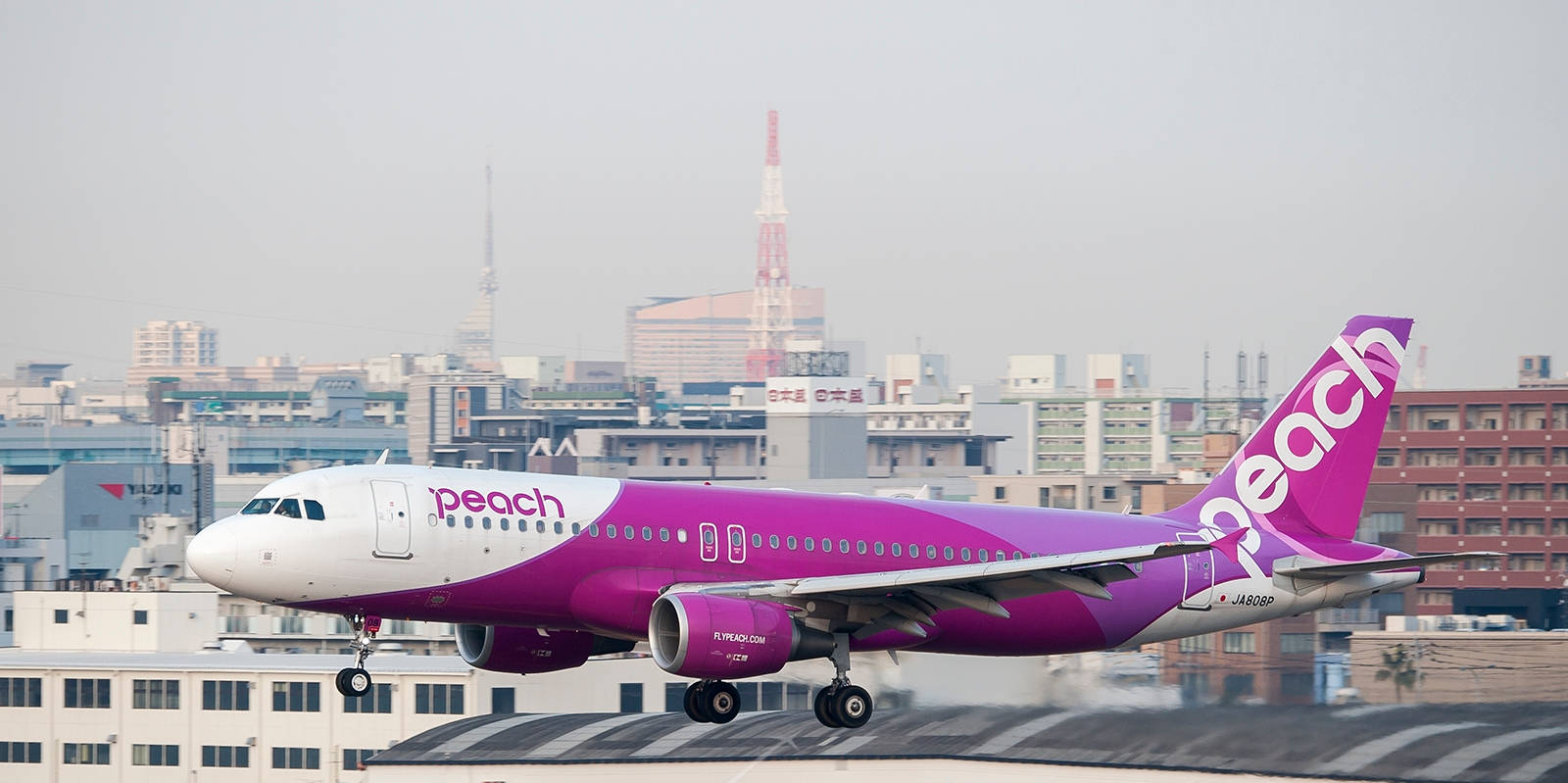 Plane 桌布 壁紙 — 一架鮮豔紫色和白色的Peach Airlines噴射機在朦朧的城市天際線背景下起飛，遠處有一座高塔。