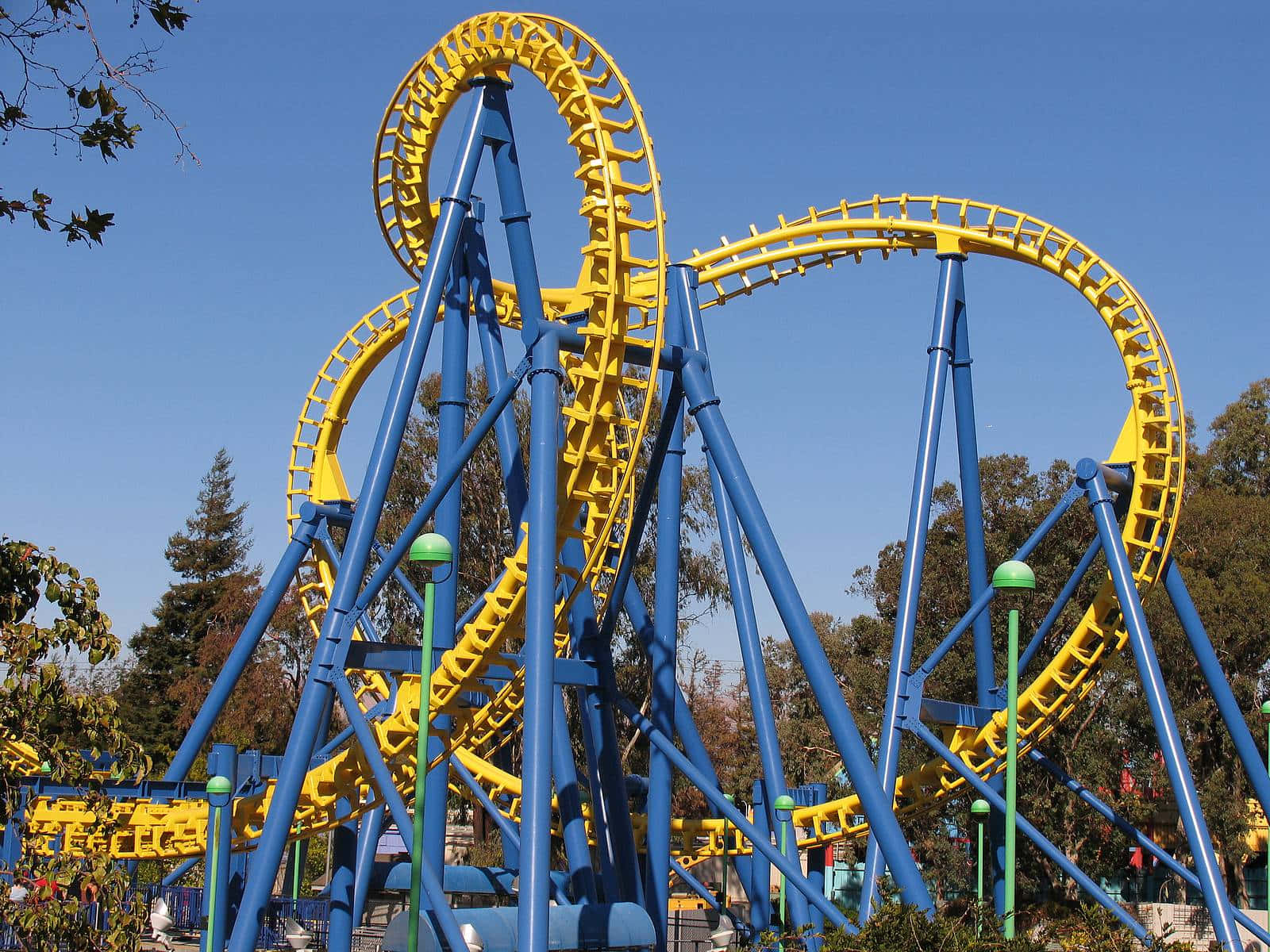 Roller Coaster 桌布 壁紙 — 一架鮮黃色和藍色的雲霄飛車在晴朗的藍天下蜿蜒曲折，背景有綠樹。