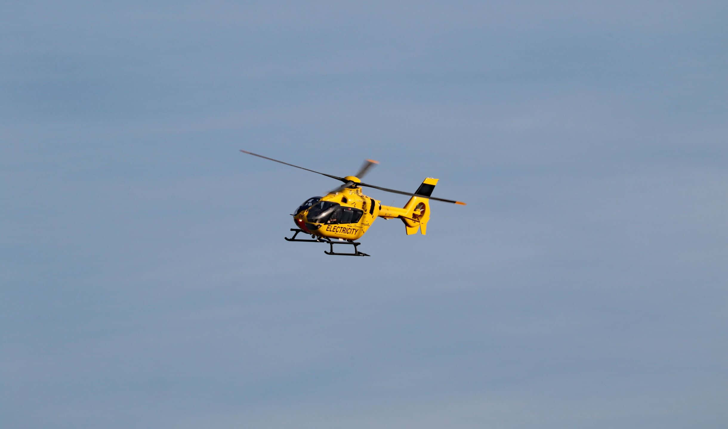 2440x1440 Helicopters 桌布 壁紙 — 一架鮮黃色的東米德蘭空中救護直升機在帶有柔和雲朵的淡藍色天空中飛行。