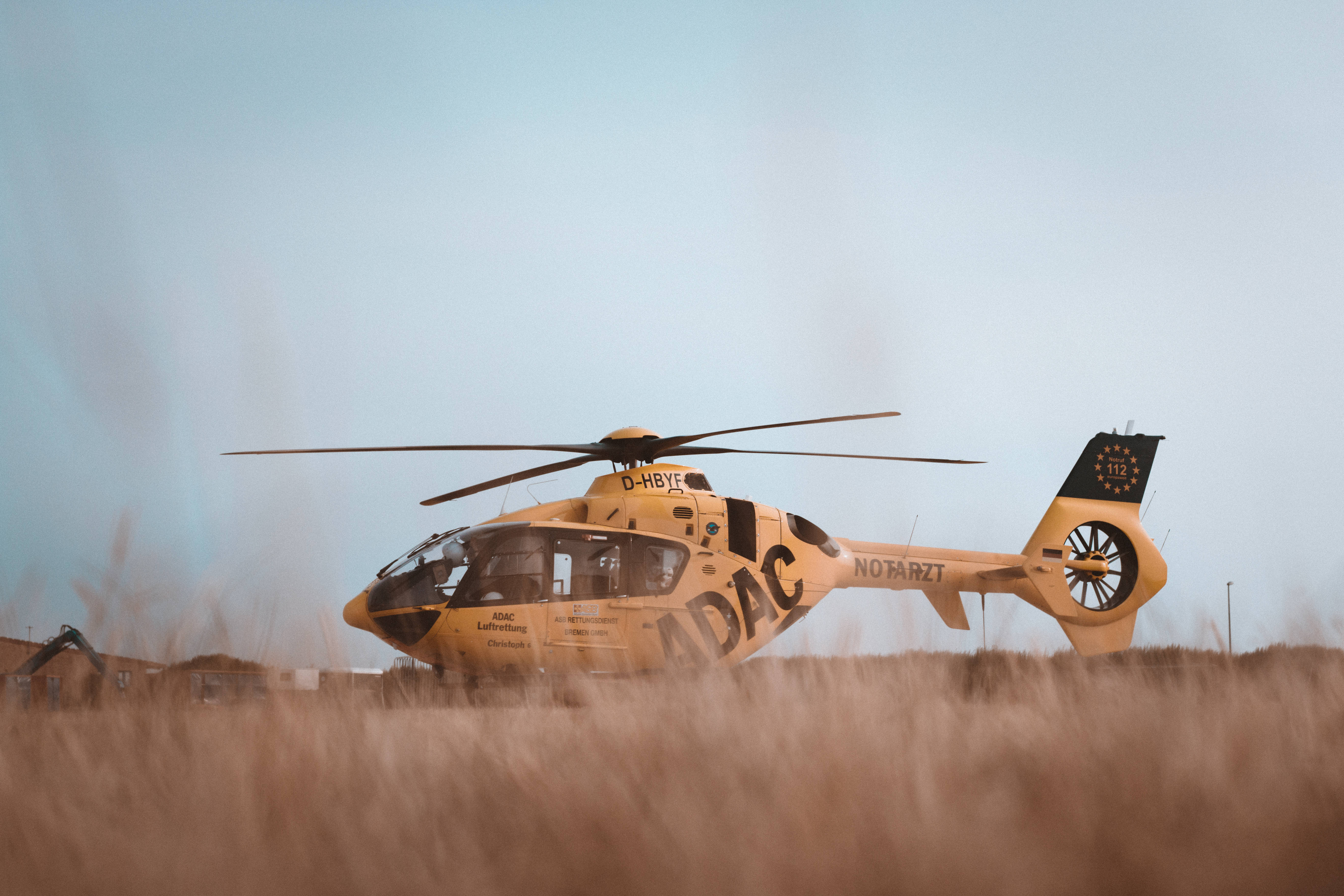 helicopter 4K 桌布 壁紙 — 一架黃色ADAC救援直升機停放在高聳乾草田野中，上方是淡藍色的天空。