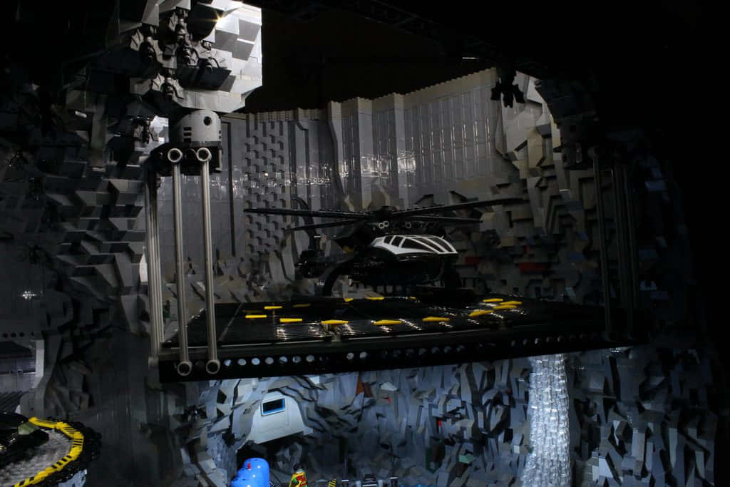 Batcave 桌布 壁紙 — 一架黑白LEGO直升機停放在一個黑暗、佈滿瓦礫的LEGO洞穴內的平台上，洞穴帶有發光的黃色點綴。