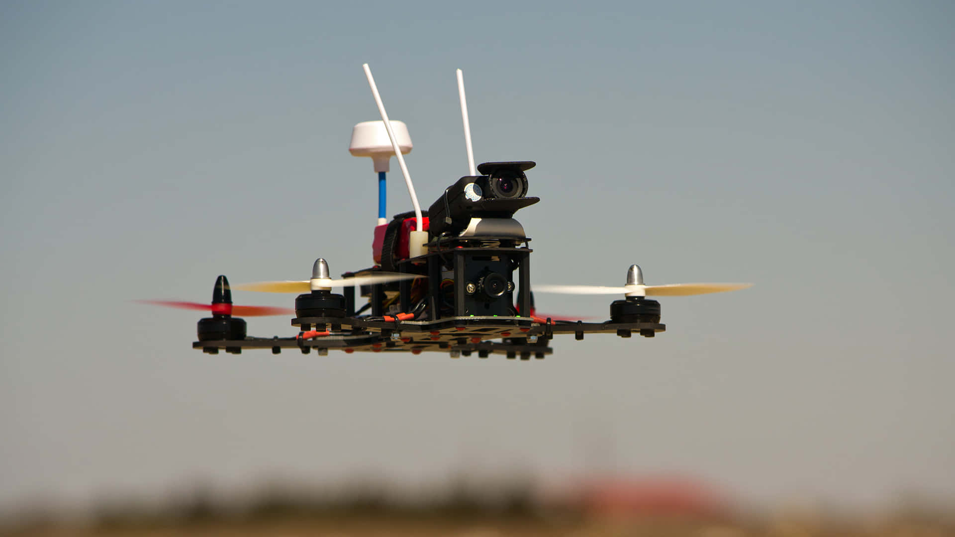 drone 桌布 壁紙 — 一架黑色四軸無人機帶著旋轉螺旋槳，在清晰藍天下盤旋，下方是模糊的地球。