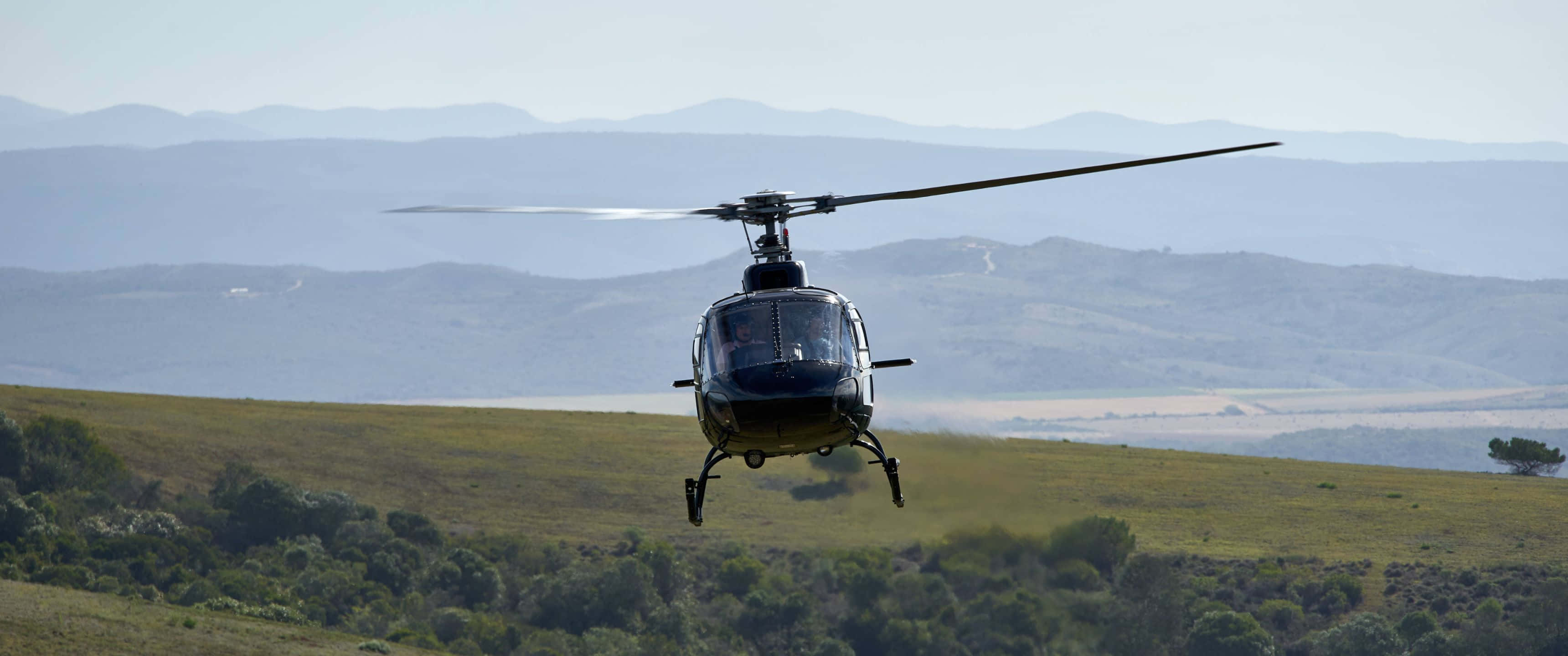 3440x1440p Helicopters 桌布 壁紙 — 一架黑色直升機低空盤旋在陽光照耀的草地上，遠處有連綿起伏的山巒，天空晴朗。