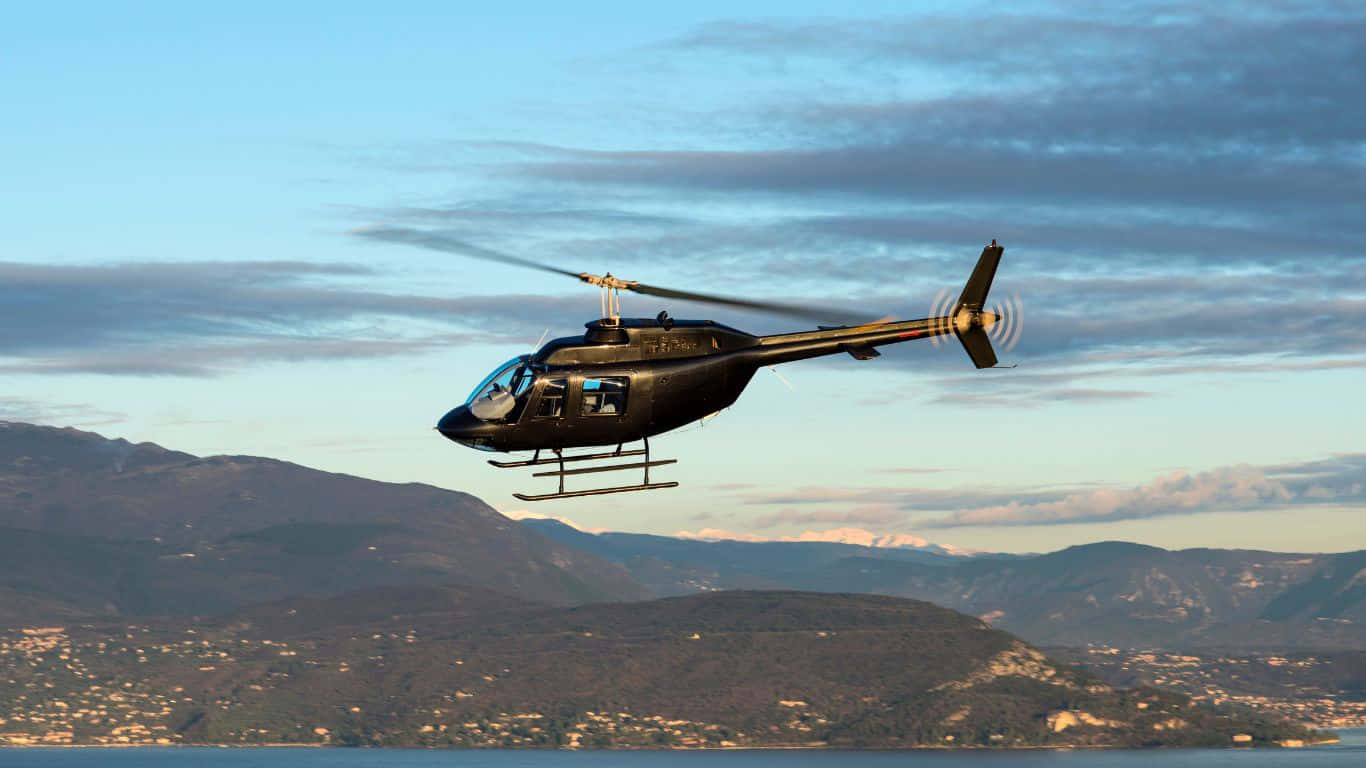1366x768 Helicopters 桌布 壁紙 — 一架黑色直升機在日落時分低飛越過多山的海岸景觀，天空有散雲。