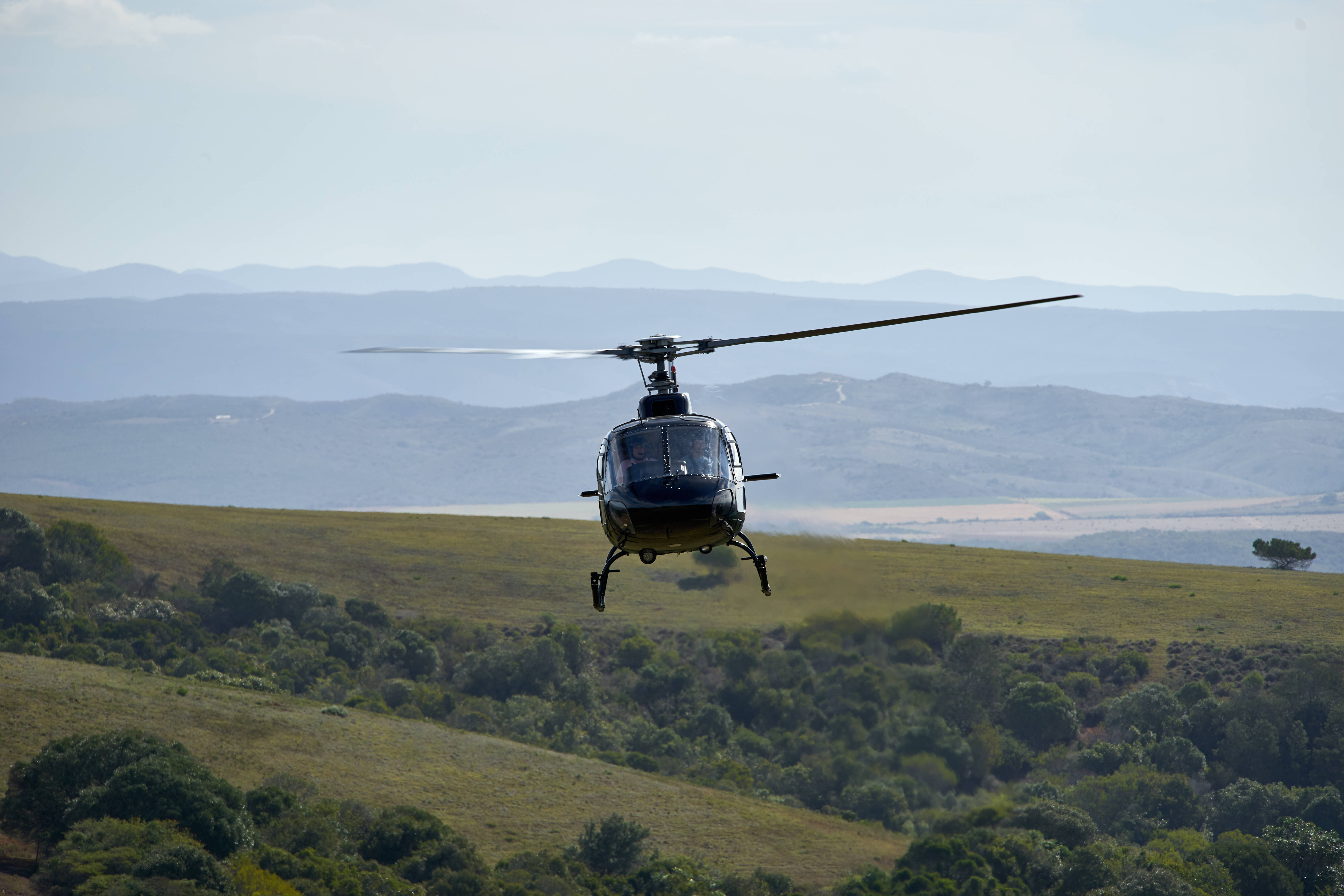 Helicopter 桌布 壁紙 — 一架黑色直升機在晴朗的天空下盤旋於起伏的綠色山丘和遠處的山脈之上。