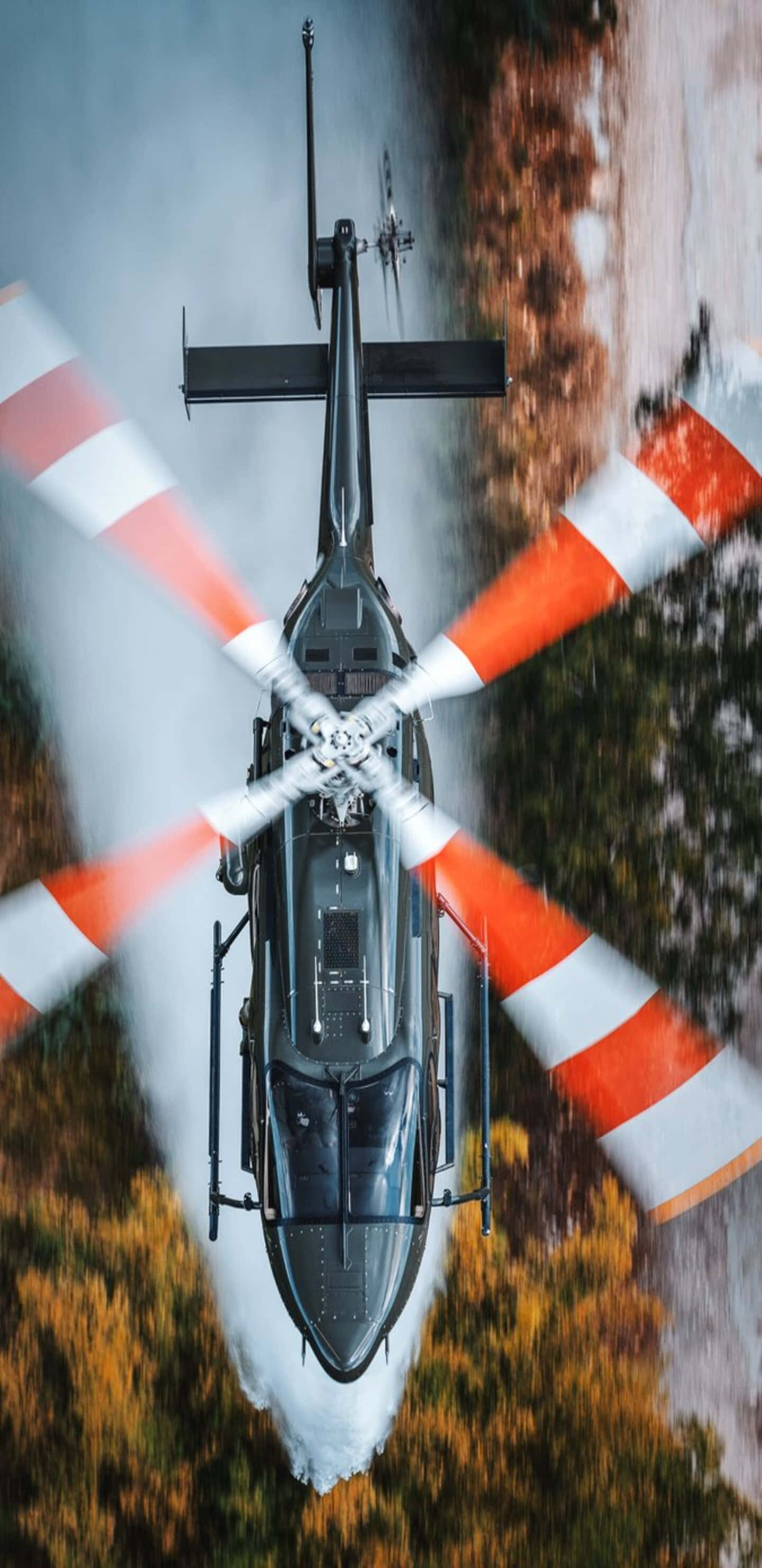 Helicopter 桌布 壁紙 — 一架黑色直升機帶有橙色和白色條紋旋翼，在動態模糊中懸浮於森林樹冠上方。