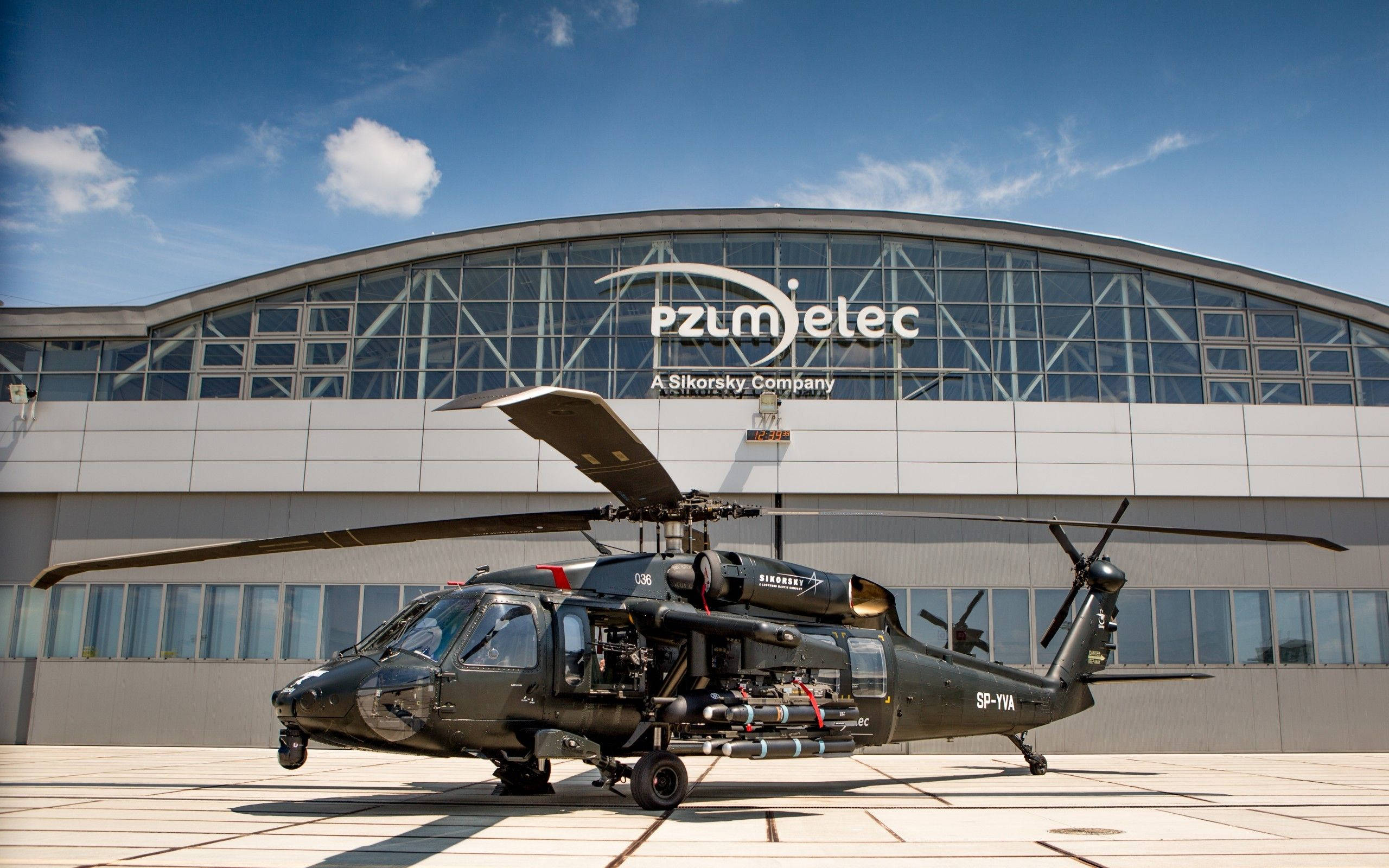 Black Hawk Helicopter 桌布 壁紙 — 一架黑色軍用直升機停在停機坪上，前方是現代化的玻璃和鋼結構機庫，背景是明亮的藍天和零星的白雲。