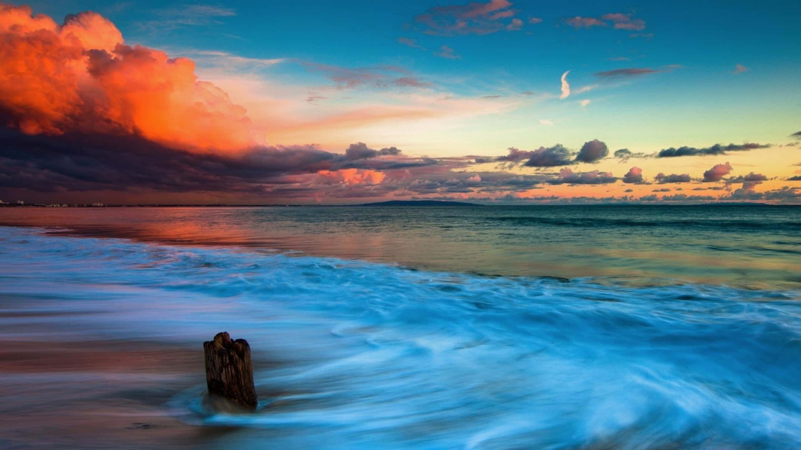 1440p Malibu 桌布 壁紙 — 一根孤獨的風化木樁矗立在泡沫翻騰的海浪中，背景是戲劇性的日落天空，有火紅的橙色雲朵和一彎新月。