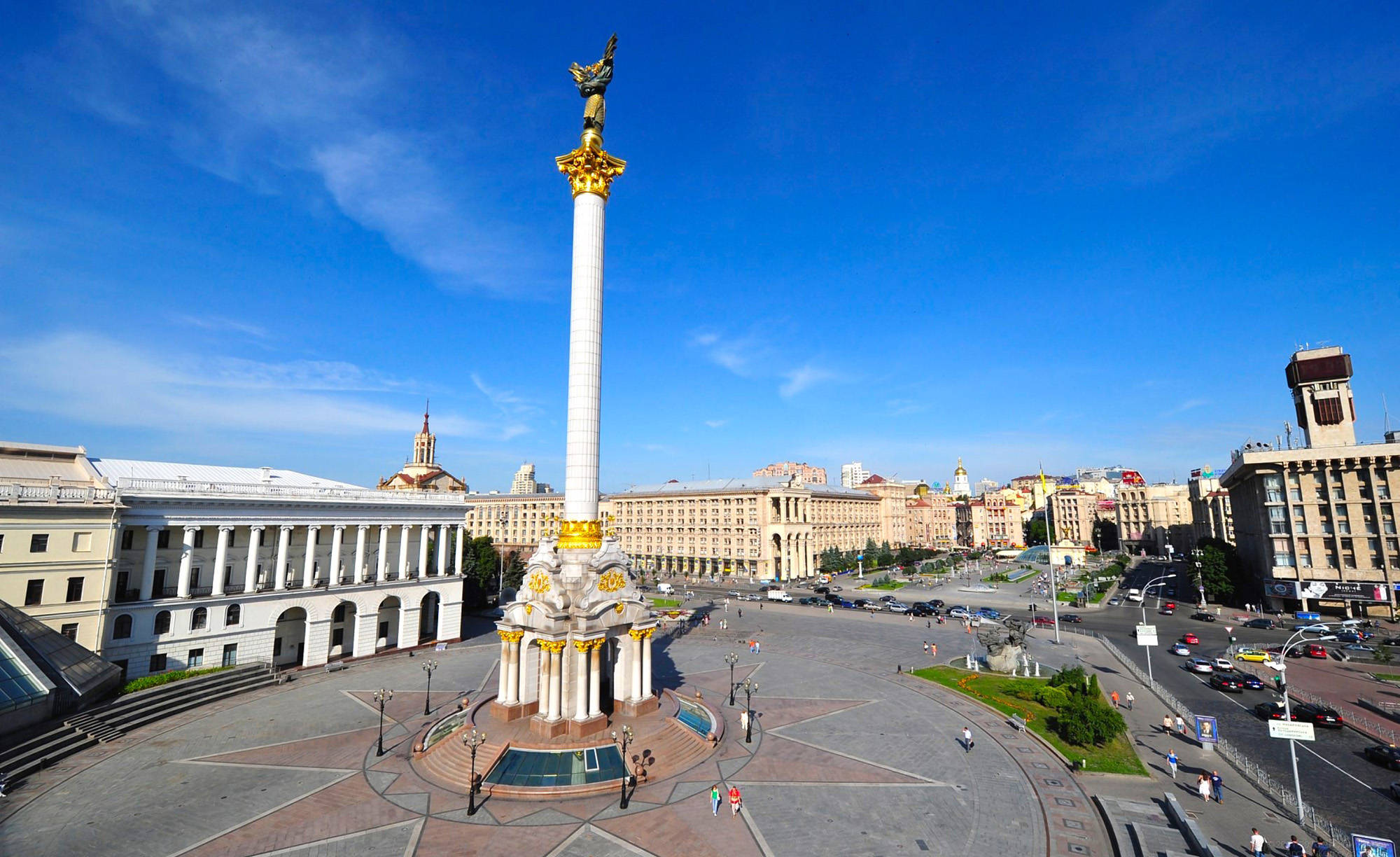 Ukraine 桌布 壁紙 — 一根巨大的白色柱子，頂端有金色天使雕像，在晴朗藍天下，聳立在陽光普照的城市廣場上，周圍是古典和現代建築。