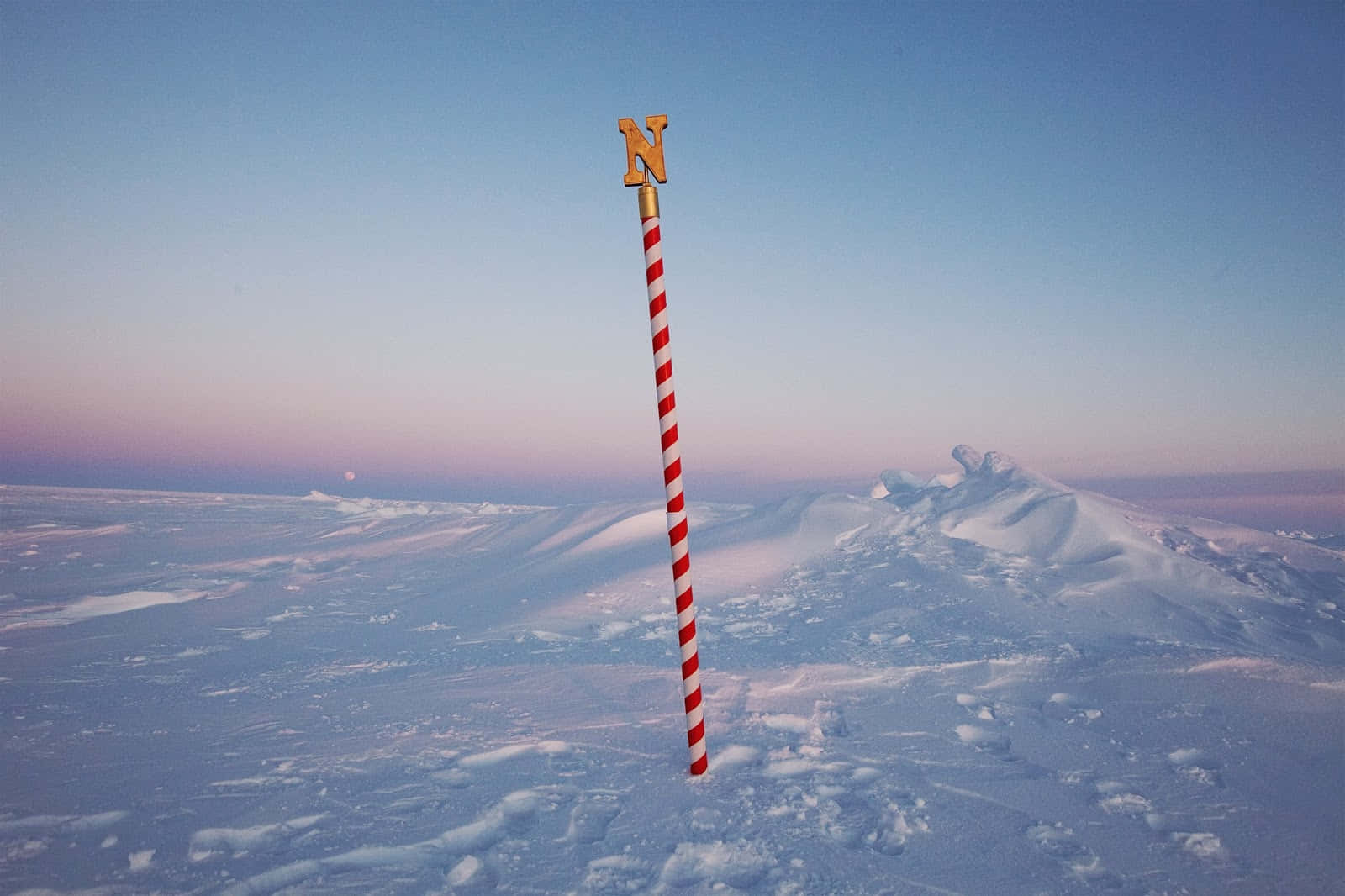 North Pole 桌布 壁紙 — 一根紅白條紋的柱子，上面有一個金色的「N」，在暮色中矗立在廣闊的雪地北極景觀中。