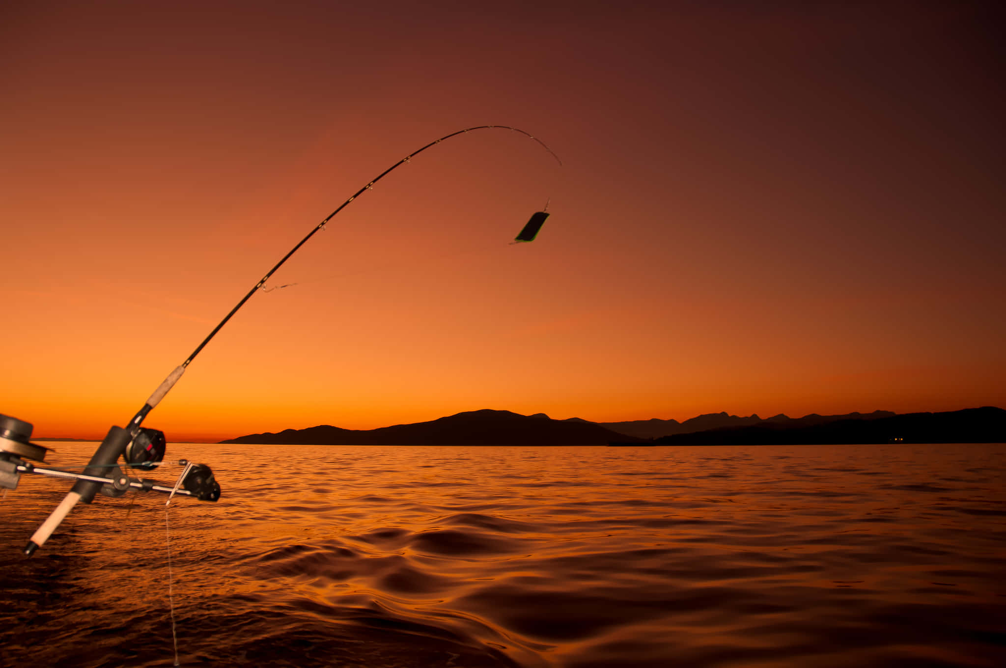 Fishing Rod 桌布 壁紙 — 一根釣竿在平靜的水面和遠山之上，對著火紅的橙色夕陽戲劇性地彎曲。