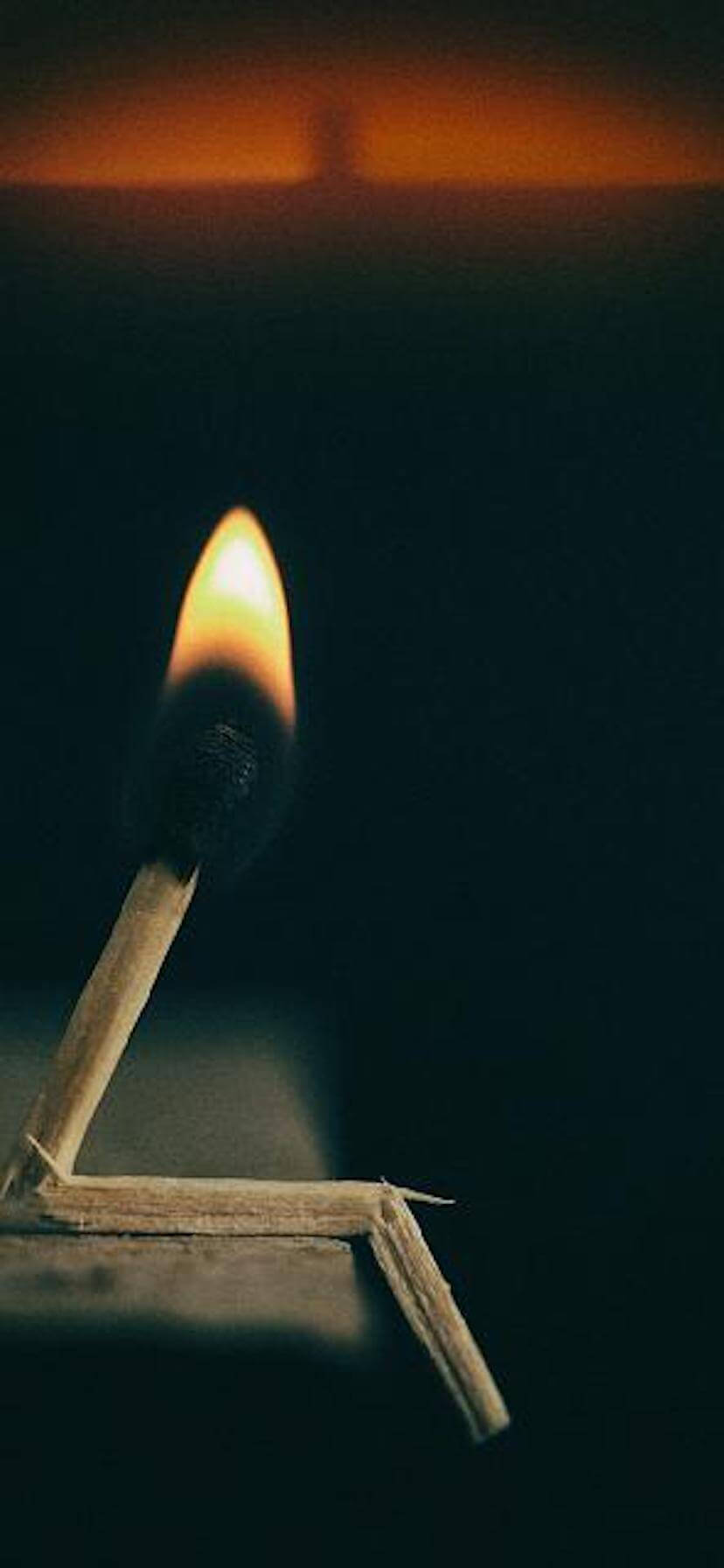 深色 iPhone 桌布 壁紙 — 一根點燃的火柴在黑暗背景上投射出溫暖的金色火焰，上方有模糊的橙色光芒。